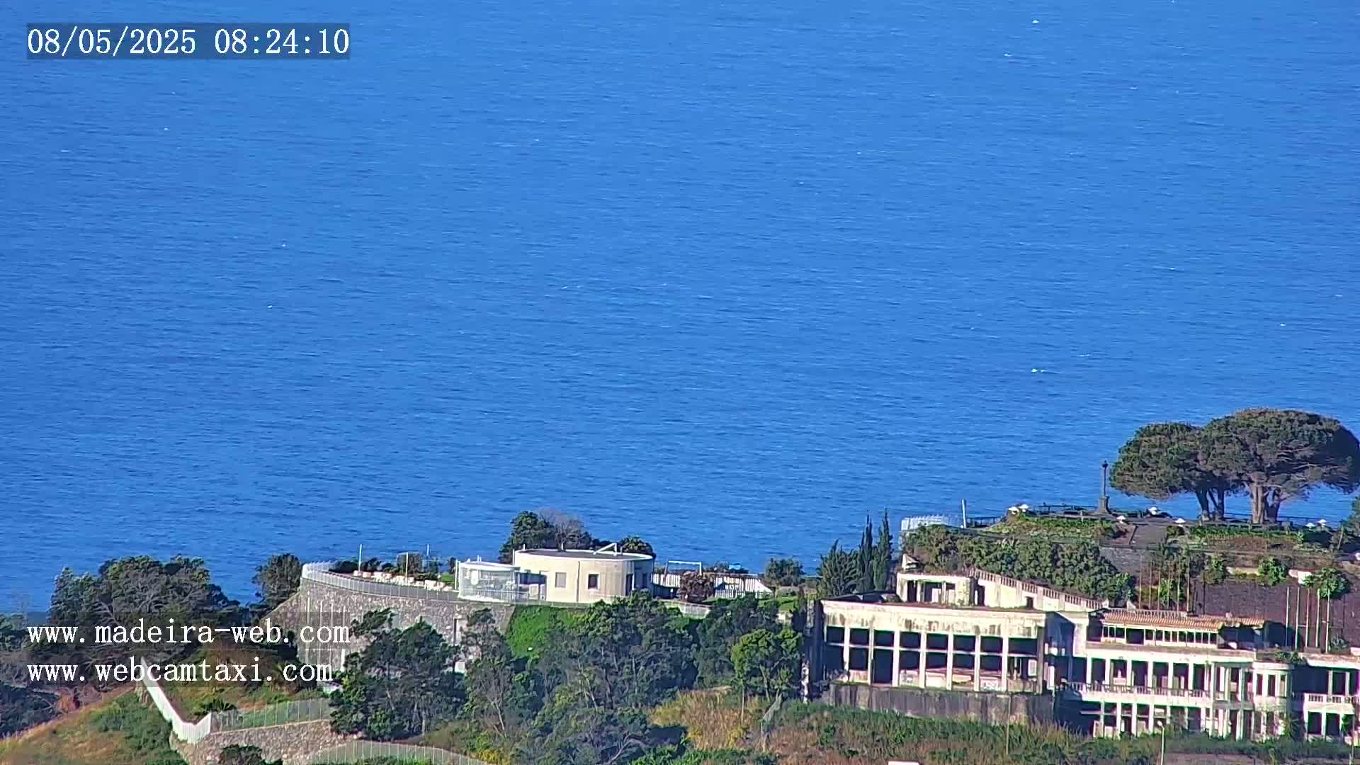 A large building complex sits on a hill overlooking a calm, blue ocean on a sunny day.