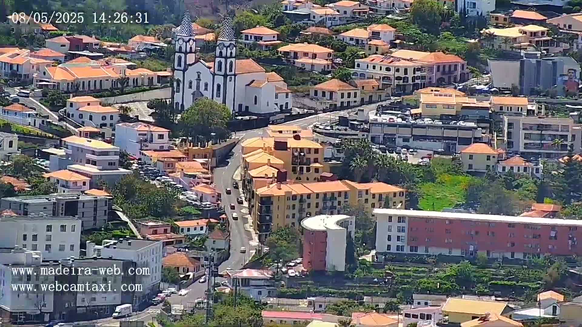 An aerial view shows a town with many low-rise buildings, a prominent church, and winding roads on a sunny day.