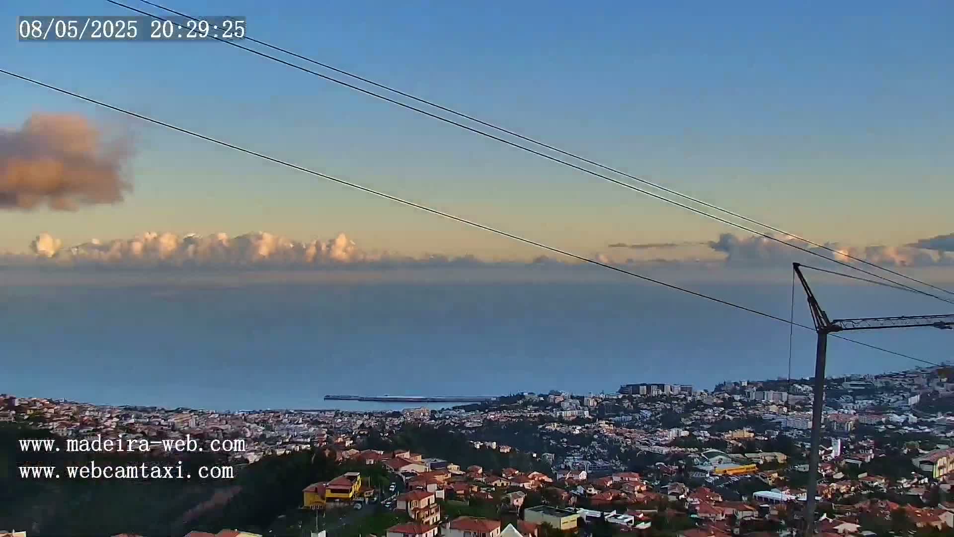 A coastal city at sunset is visible beneath a partly cloudy sky, with power lines and a crane in the foreground.