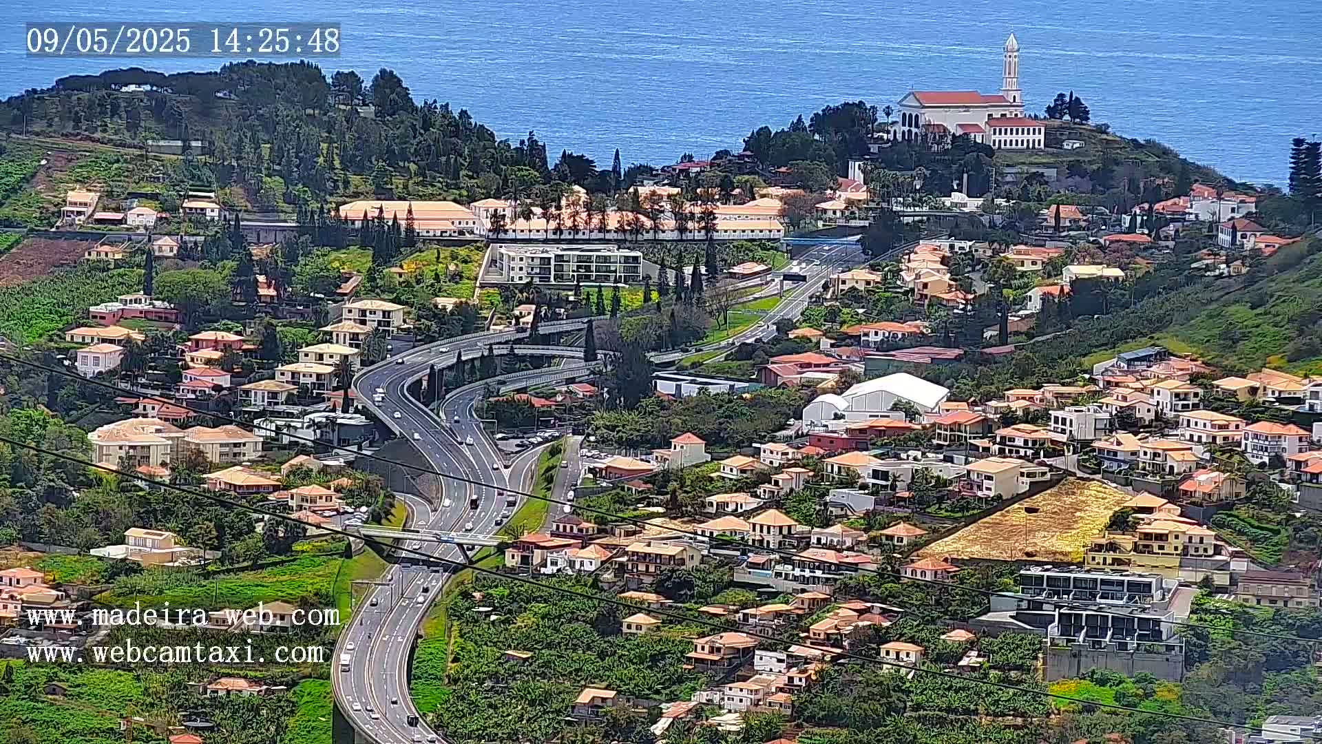 A coastal hillside town with a white church and a multi-lane highway, all under sunny skies and overlooking a blue ocean.