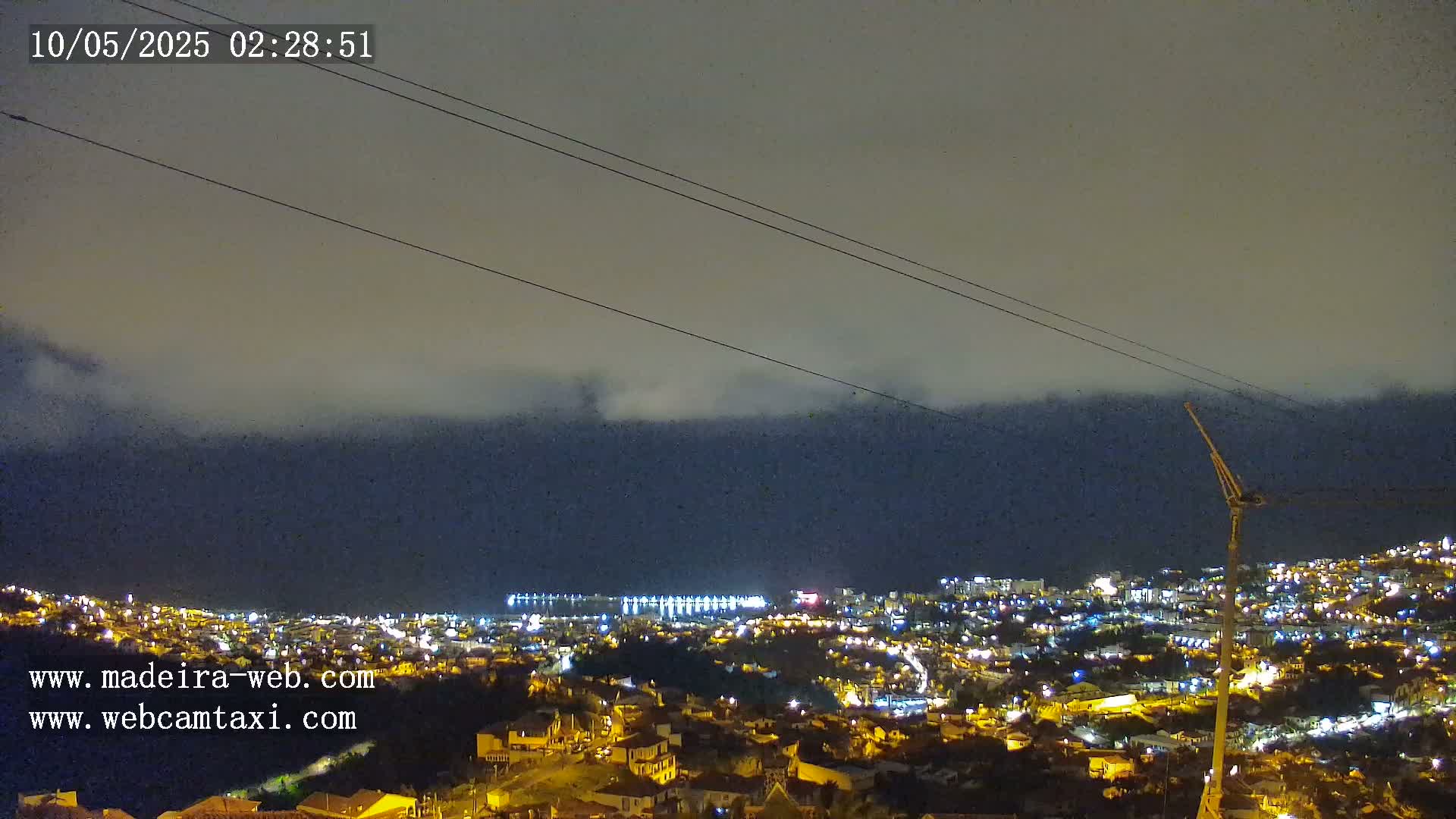 A nighttime view of a coastal town illuminated by lights, with a hazy or foggy atmosphere and power lines in the foreground.