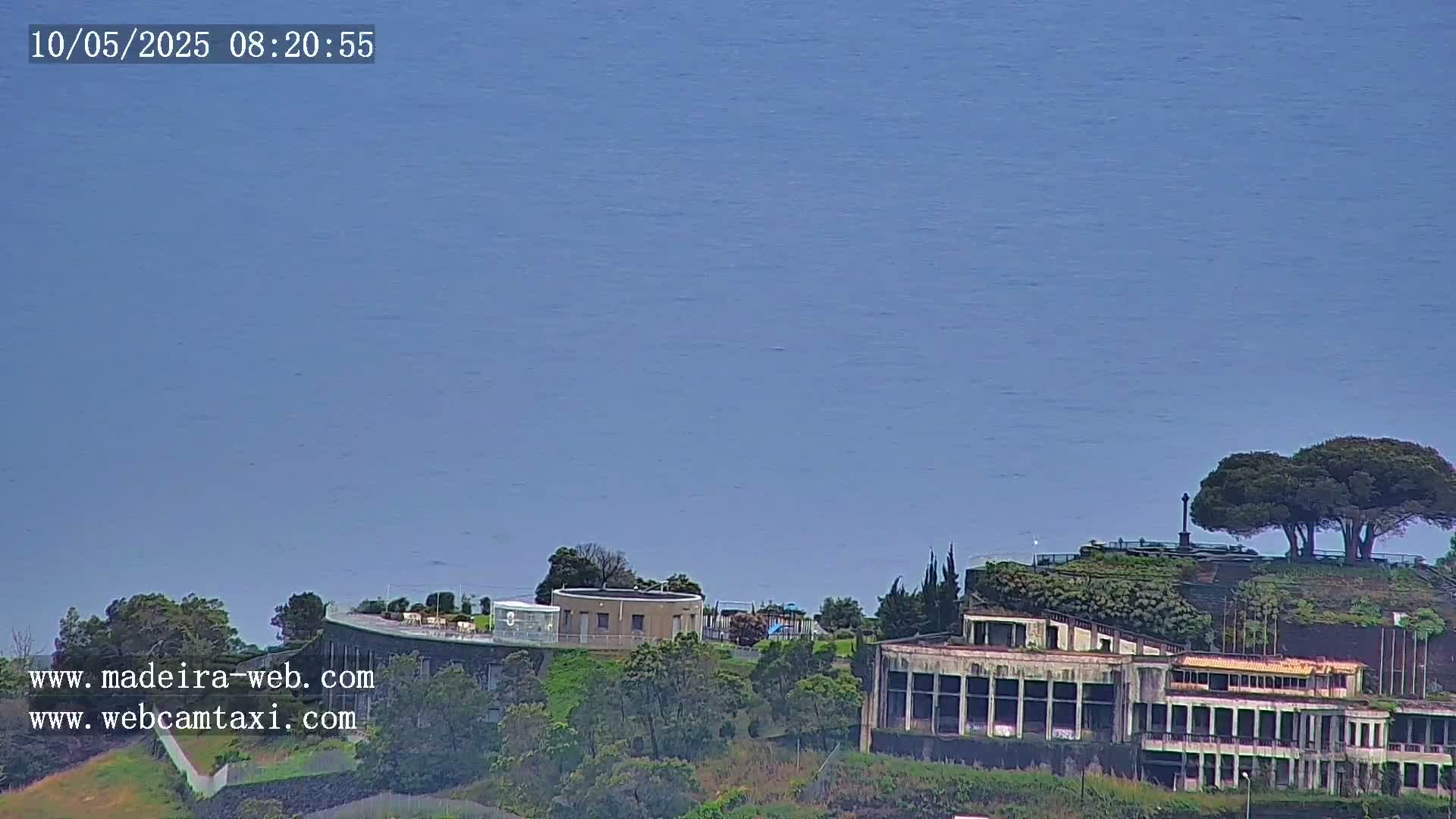 A large building complex sits on a hill overlooking a calm, blue ocean under a clear sky.