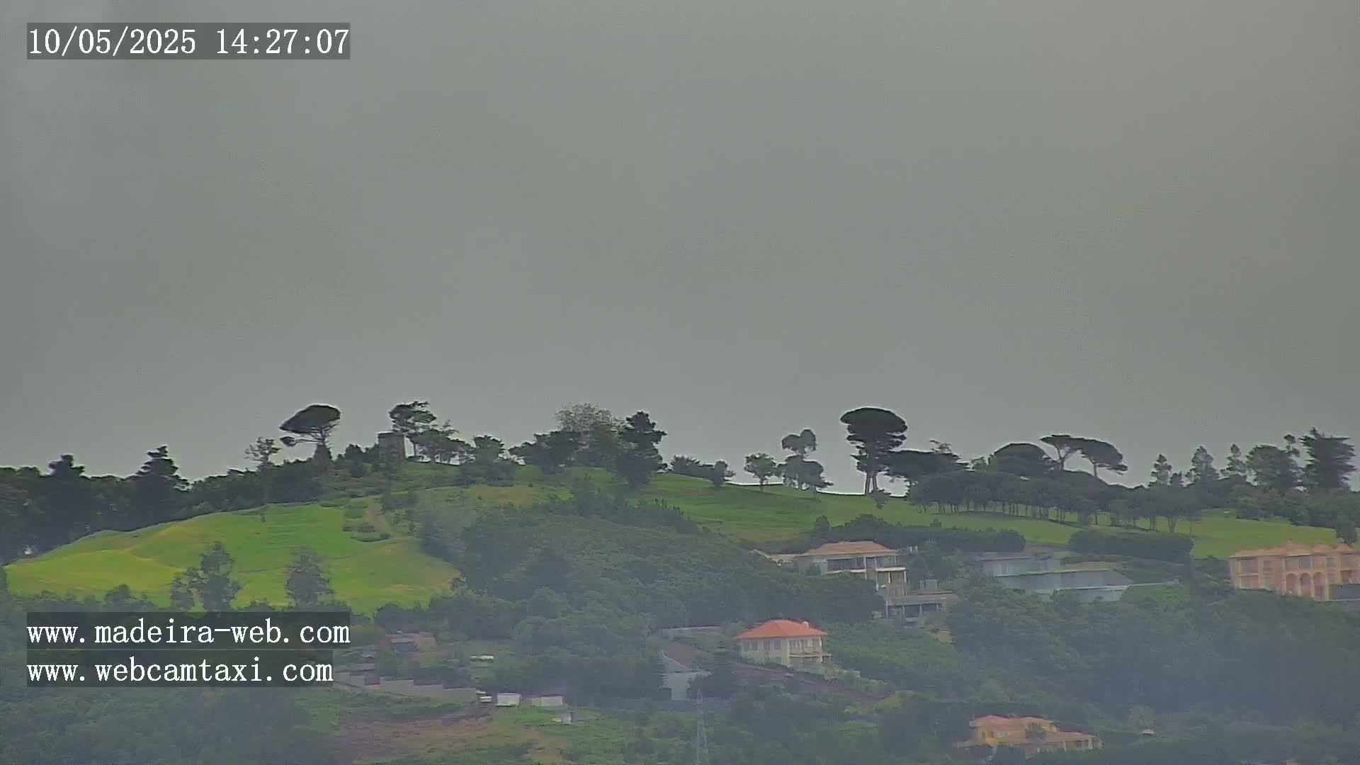 A hazy, overcast sky hangs over a green hillside dotted with trees and houses.