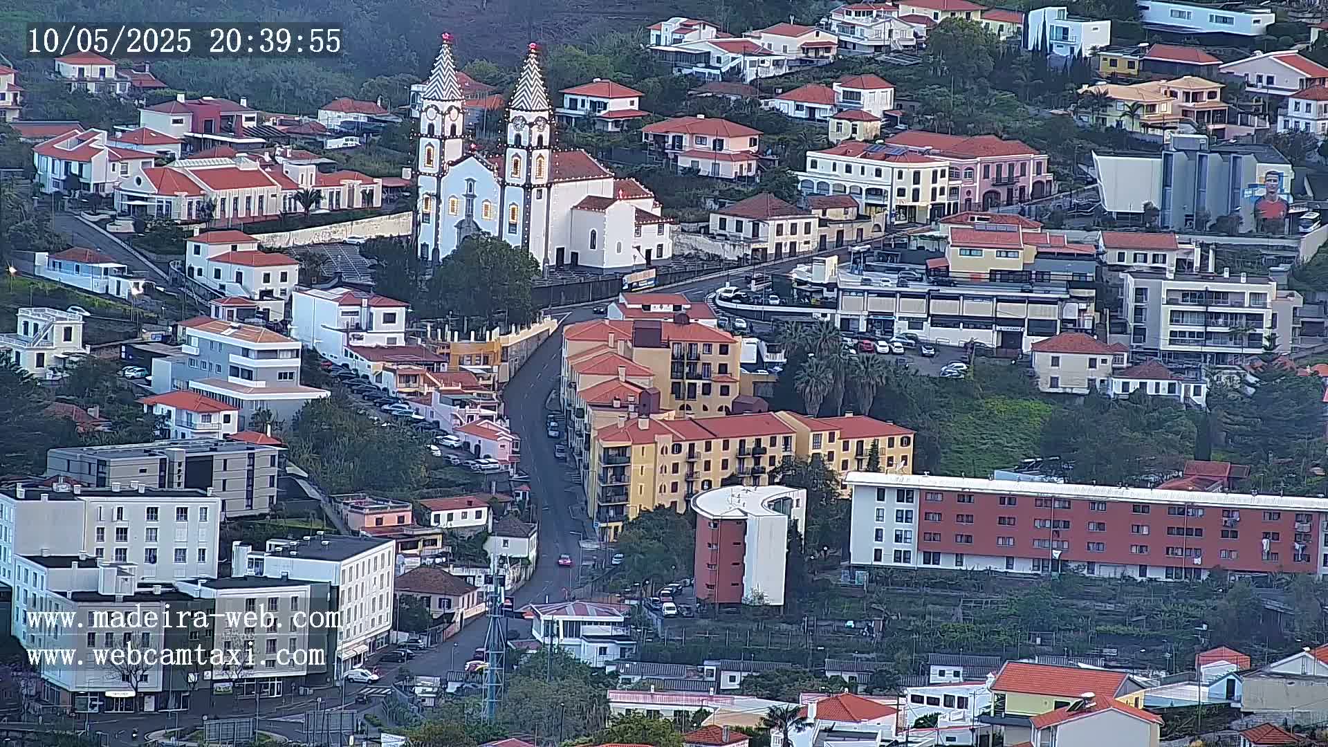 An aerial view of a town at dusk shows a church with twin towers, numerous residential buildings, and winding streets.