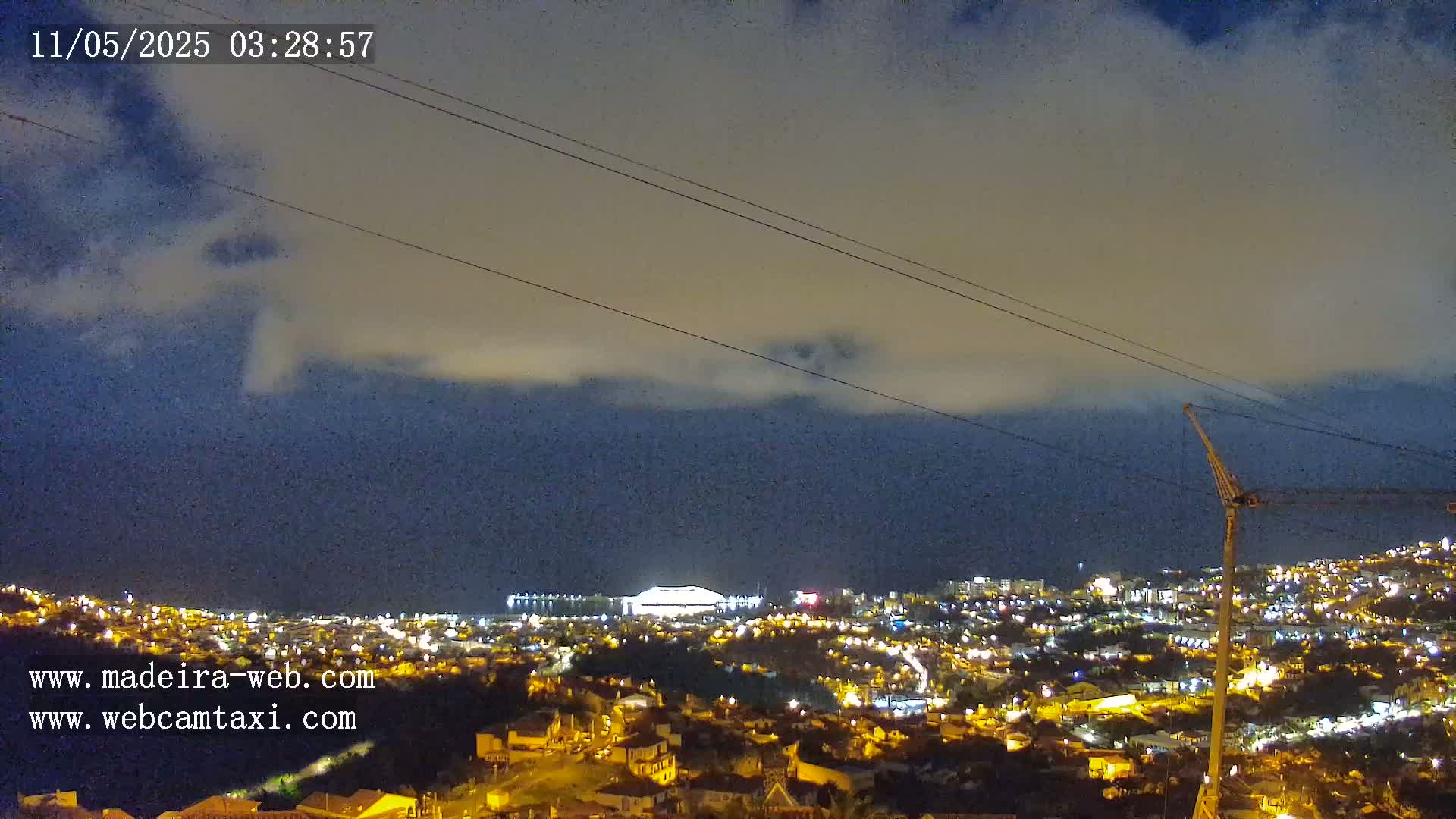 A nighttime view of a city illuminated by lights, partially obscured by clouds under a dark sky, with a construction crane visible in the bottom right corner.