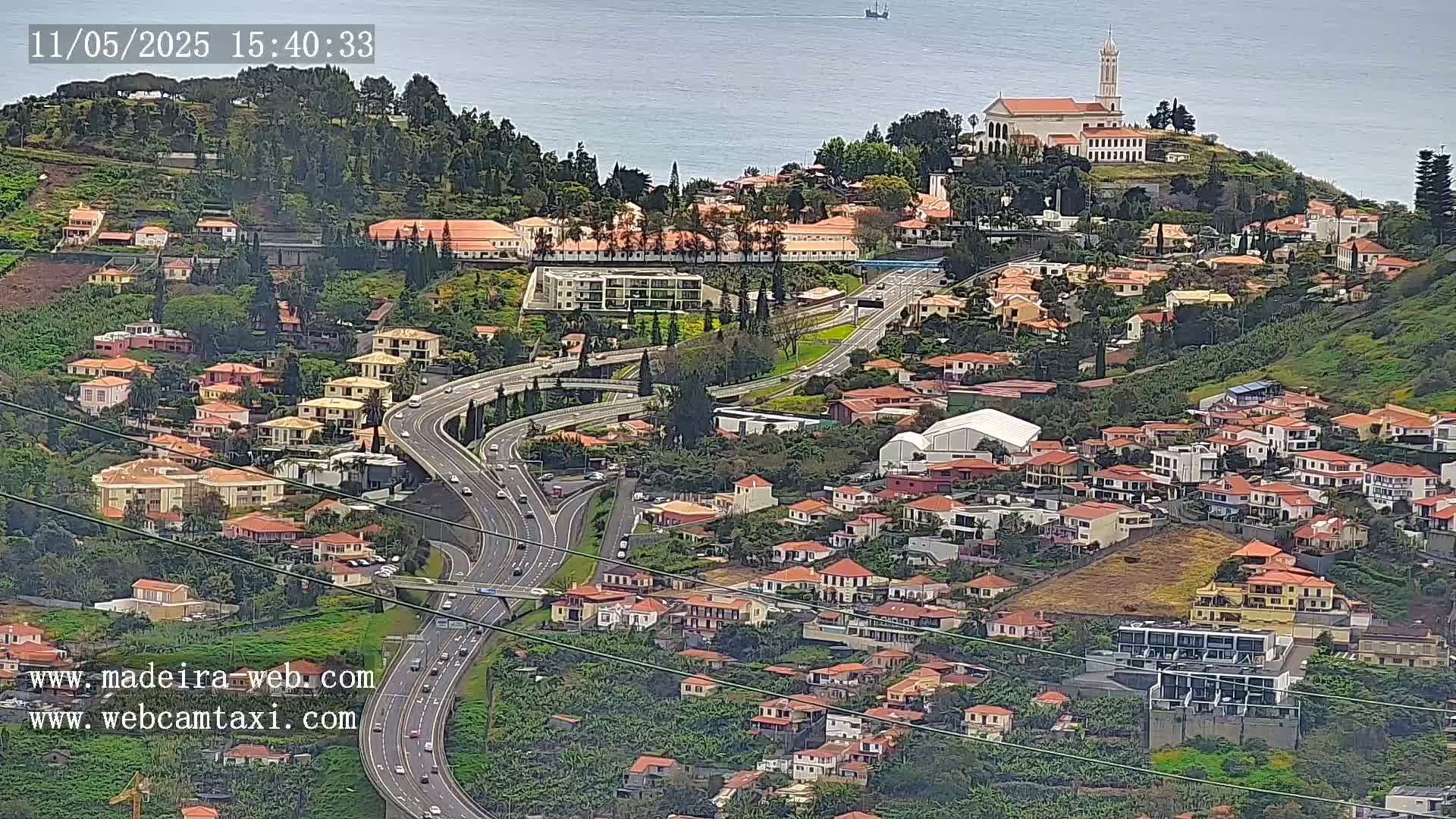 An aerial view shows a coastal town with a church atop a hill, many houses, and a multi-lane highway weaving through the landscape on a partly cloudy day.