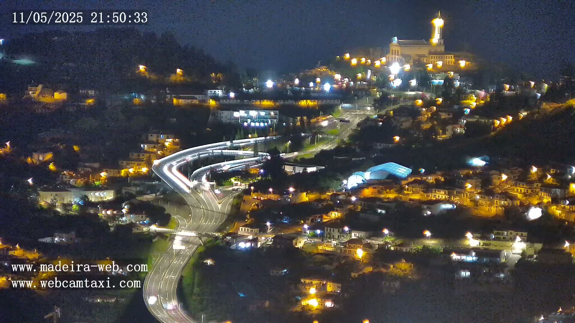 A nighttime aerial view of a town built on a hillside, illuminated by streetlights, with a church atop the hill and a highway interchange weaving through the lower parts of the town.