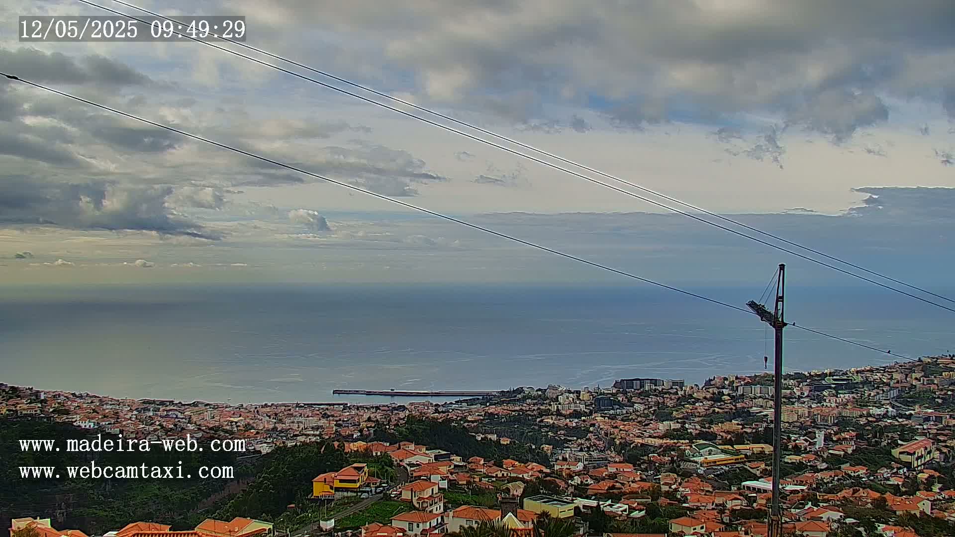 A coastal town with red-roofed houses stretches below a cloudy sky, overlooking a calm ocean.
