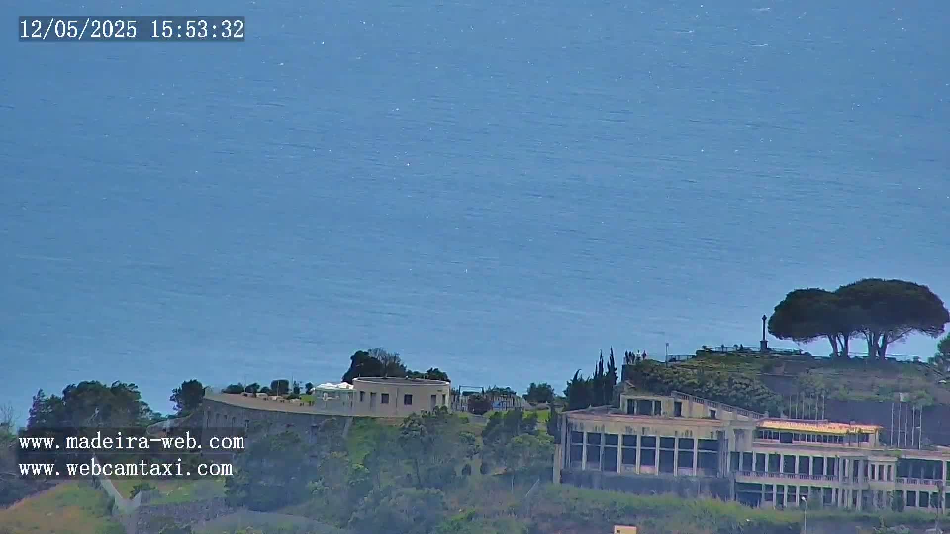 A large building complex sits atop a hill overlooking a calm, blue ocean under a clear sky.