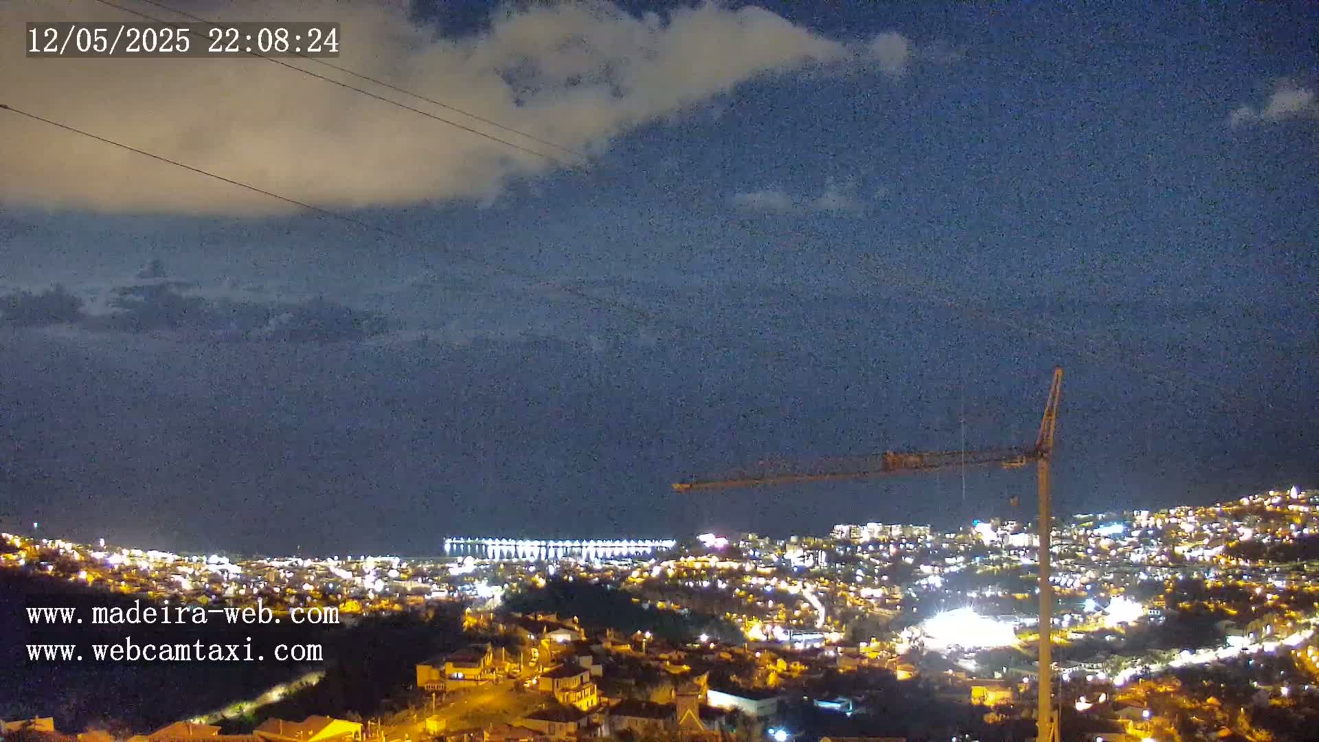 A nighttime view of a city illuminated by lights, partially obscured by clouds, with a construction crane visible in the foreground and a body of water in the distance.