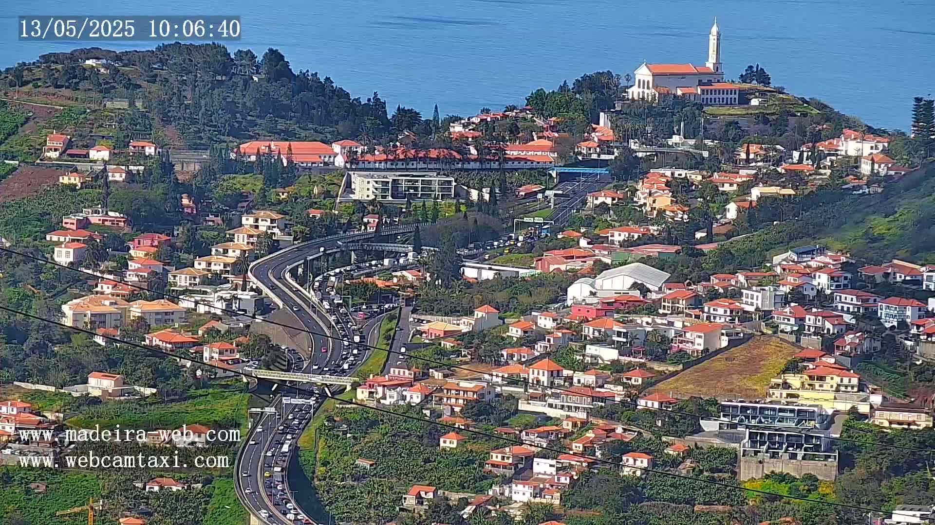 A hillside town with red-roofed houses and a church, overlooks a busy highway with heavy traffic, all under a clear sunny sky and next to a body of blue water.
