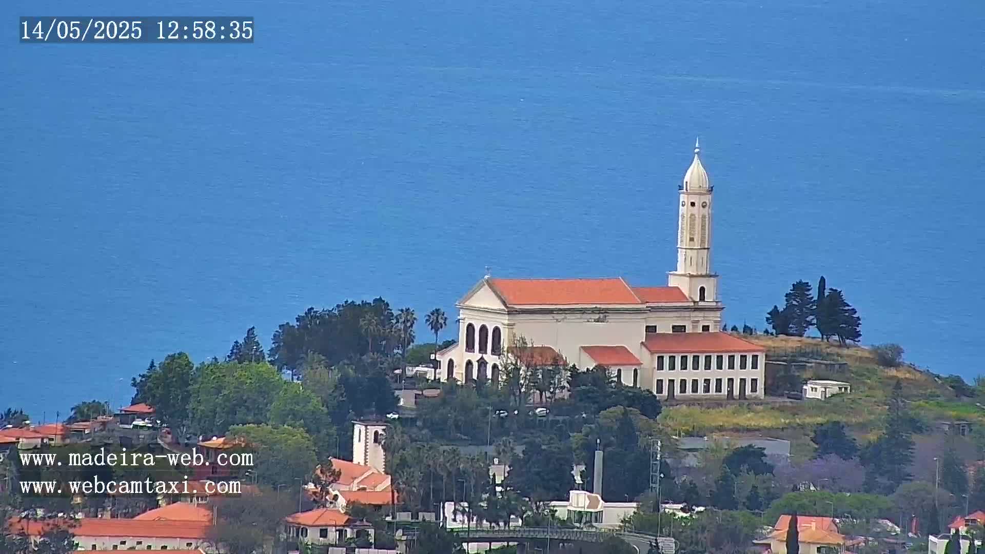 A white church with a tall tower sits atop a small hill overlooking a calm blue ocean on a clear day.