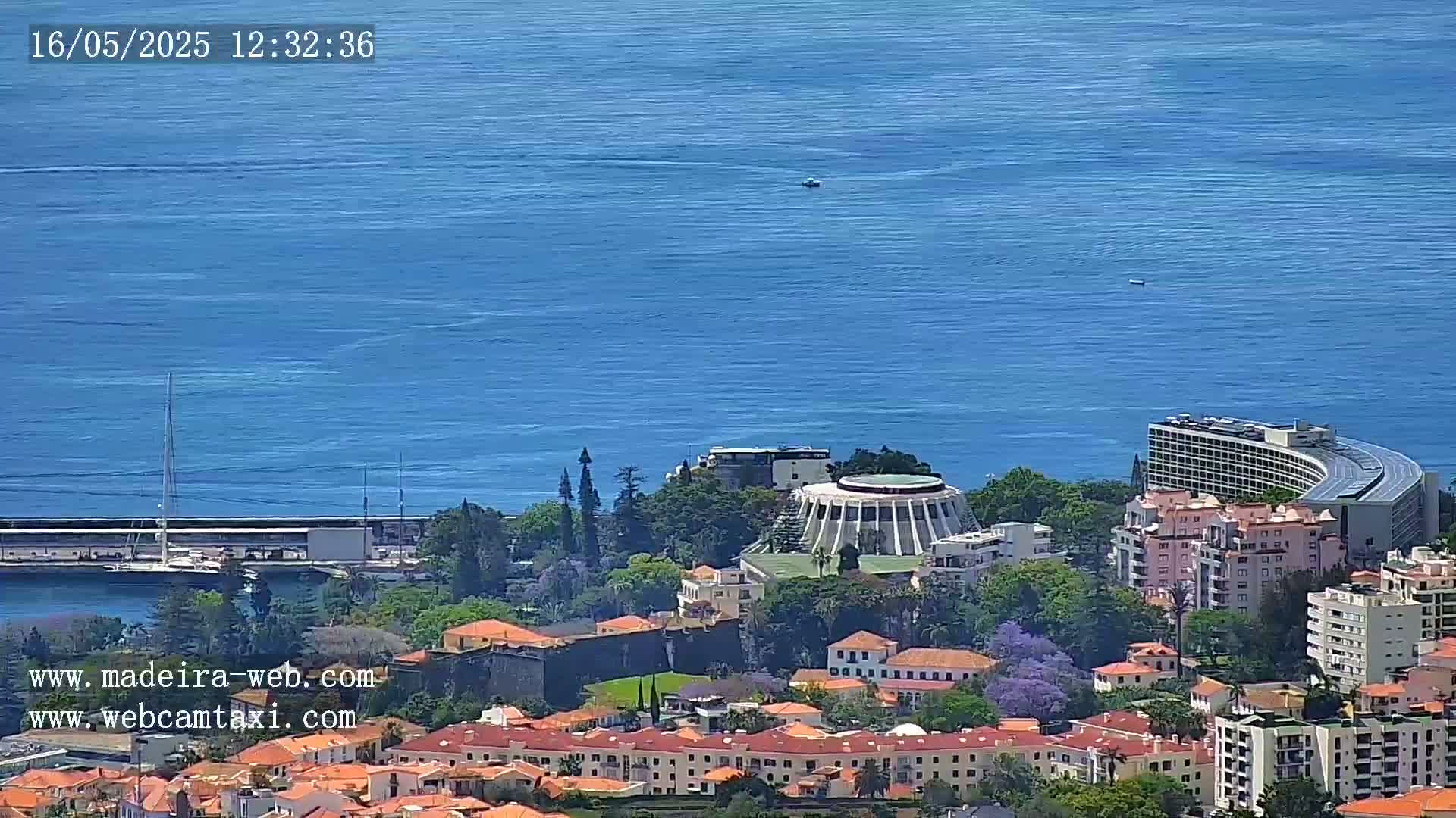 A coastal city with red-roofed buildings, lush green vegetation, and a large circular structure is visible on a sunny day, overlooking a calm blue ocean.