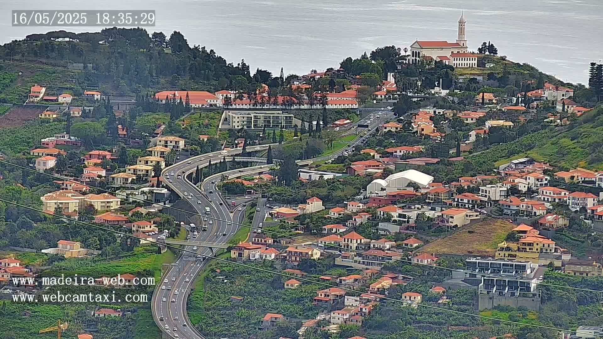 An aerial view shows a hillside town with red-roofed houses, a church on a hilltop, and a winding highway below, under an overcast sky.