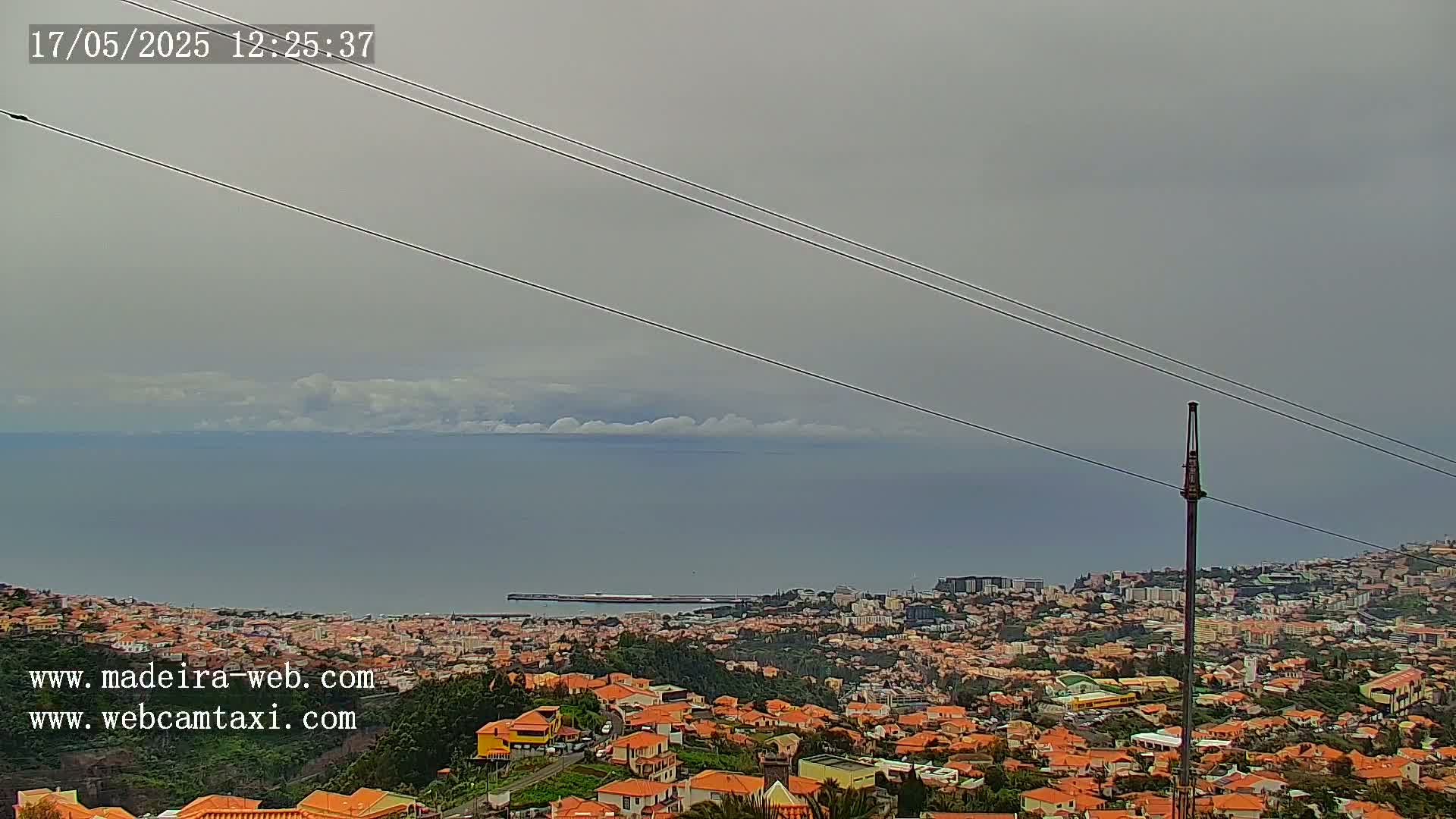 A coastal city with orange-roofed buildings stretches toward a calm ocean under an overcast sky.
