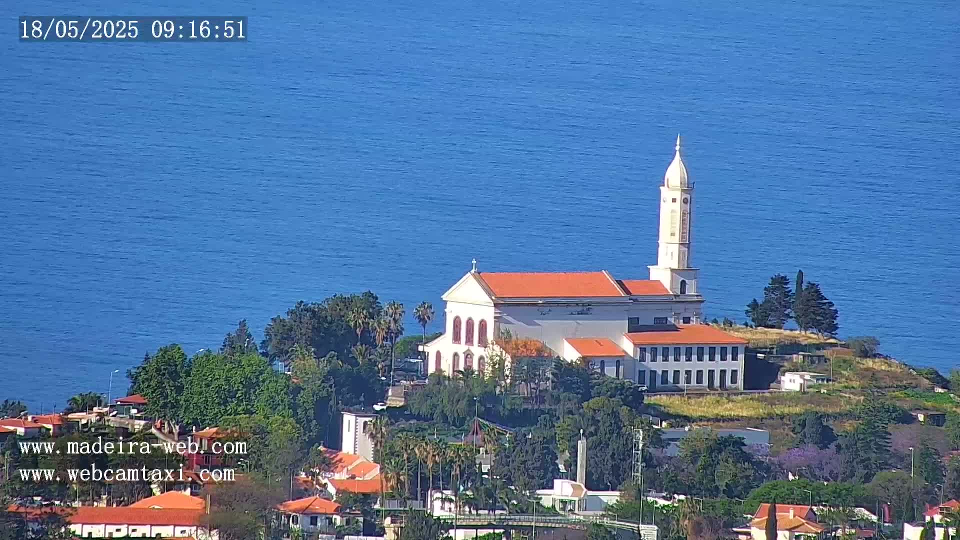 A white church with an orange roof sits on a hill overlooking a calm blue ocean under a clear sky.