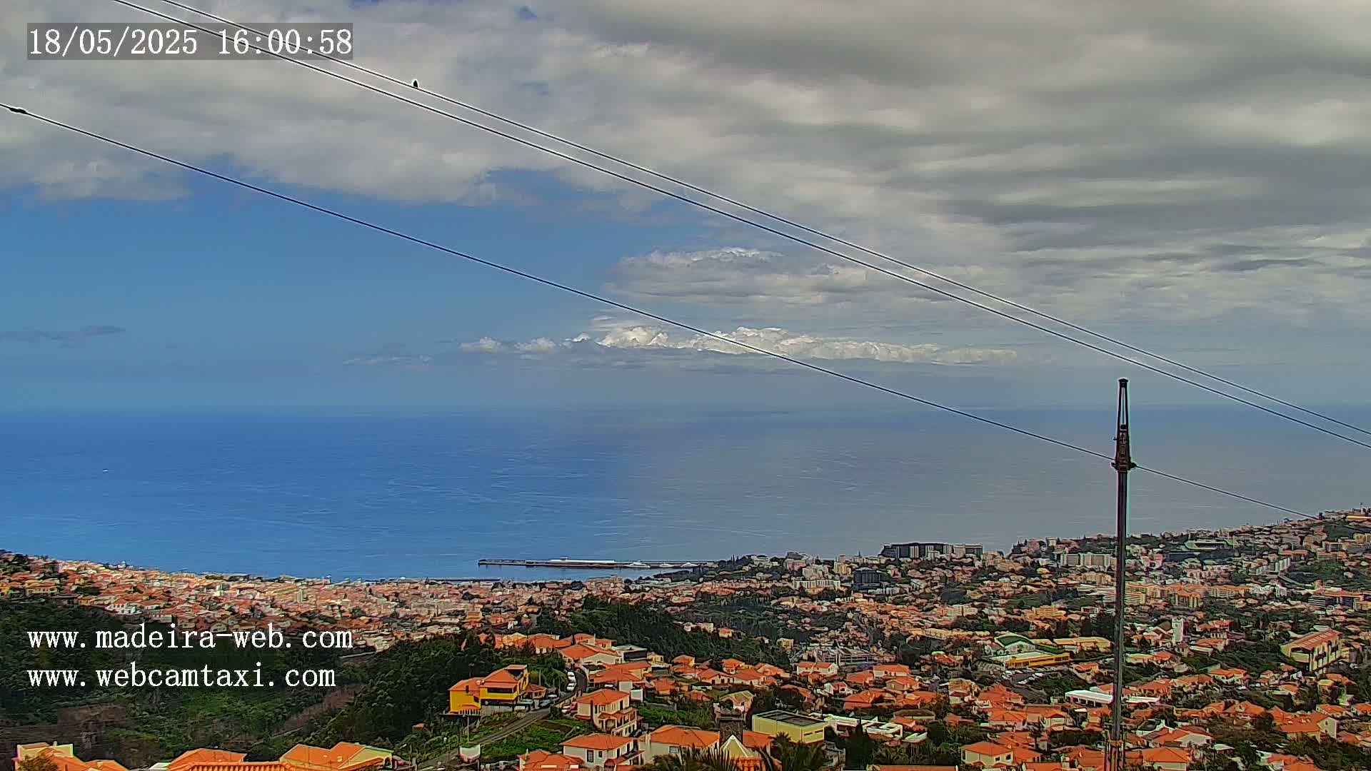 A coastal city with red-roofed buildings stretches towards a calm ocean under a partly cloudy sky, with distant mountains visible on the horizon.
