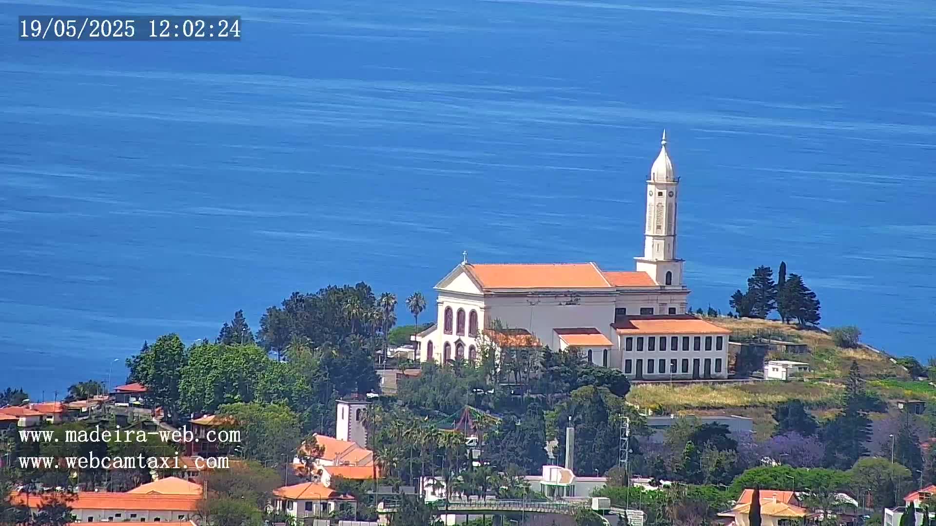 A white church with an orange roof sits atop a small hill overlooking a calm blue ocean on a sunny day.
