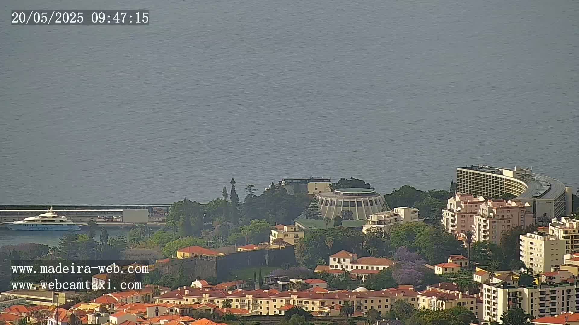 A high-angle, long shot shows a coastal city on a partly cloudy day, with a large body of calm water in the background.