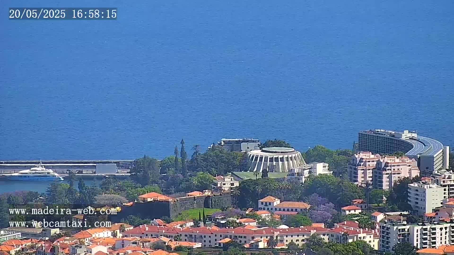 A coastal city with red-roofed buildings, a large circular building, and a curved hotel overlooks a calm blue ocean on a sunny day.