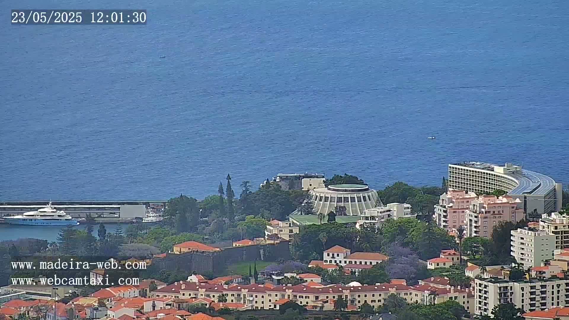 A coastal town with numerous buildings, including a large hotel and a circular structure, overlooks a calm blue ocean on a clear day.