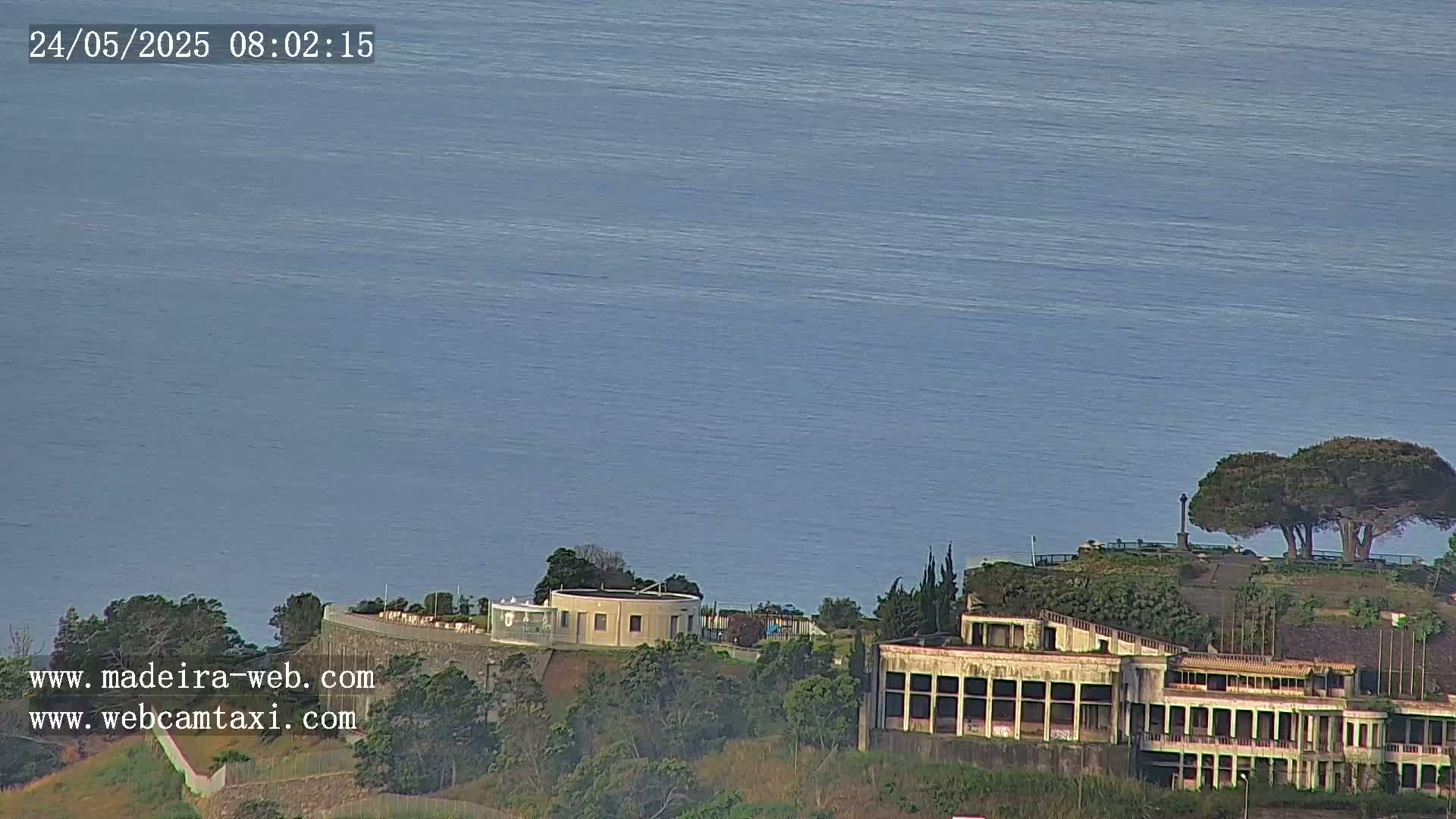 A large building complex sits on a hillside overlooking a calm, blue ocean under a clear sky.