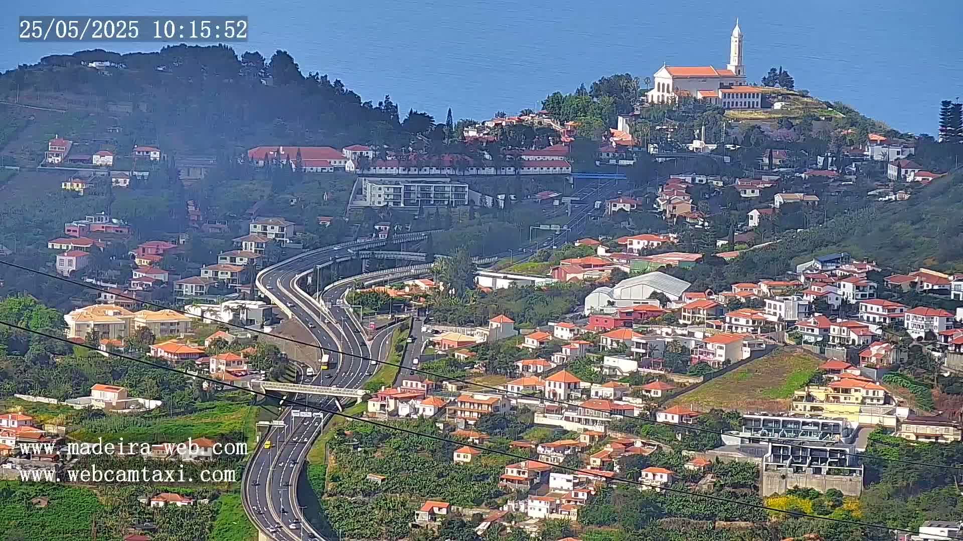A hillside town with red-roofed houses and a church on a hilltop overlooks a highway curving through the landscape on a sunny day.