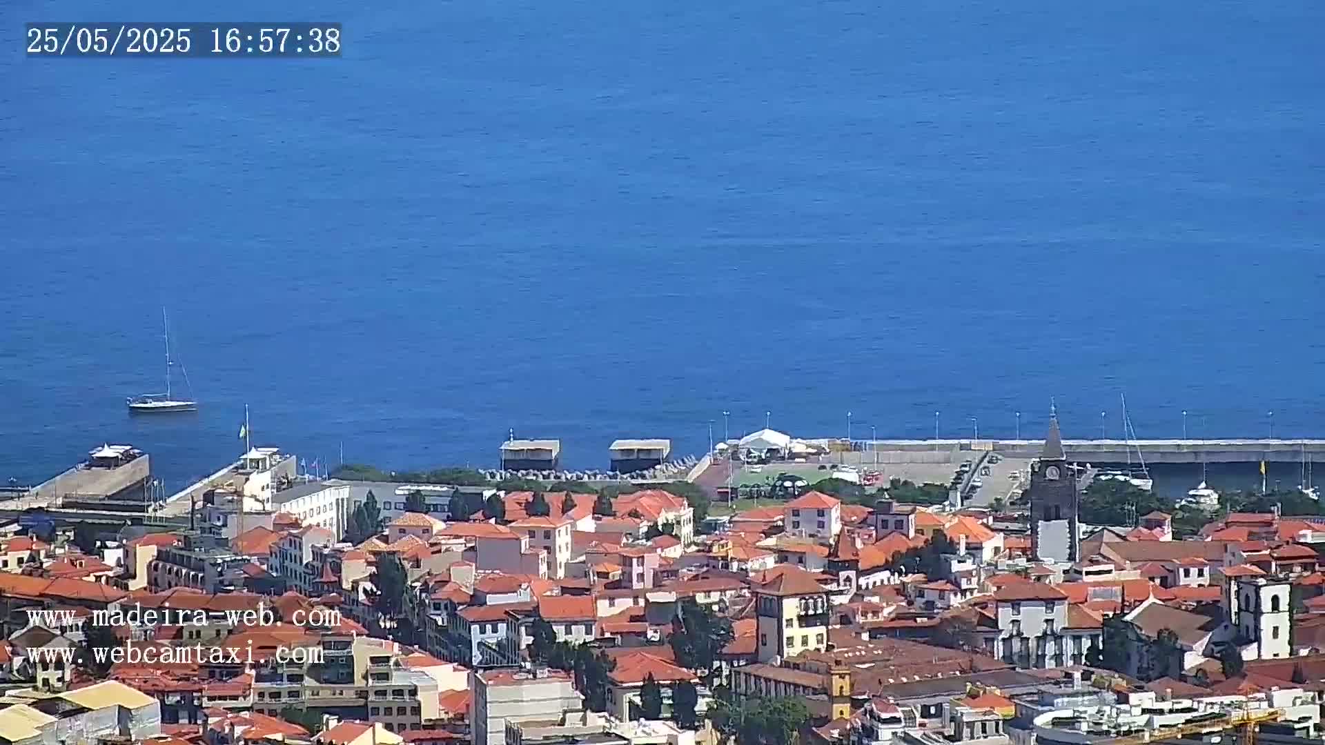 A coastal town with red-roofed buildings, a harbor, and a small sailboat is visible under a clear, sunny sky.