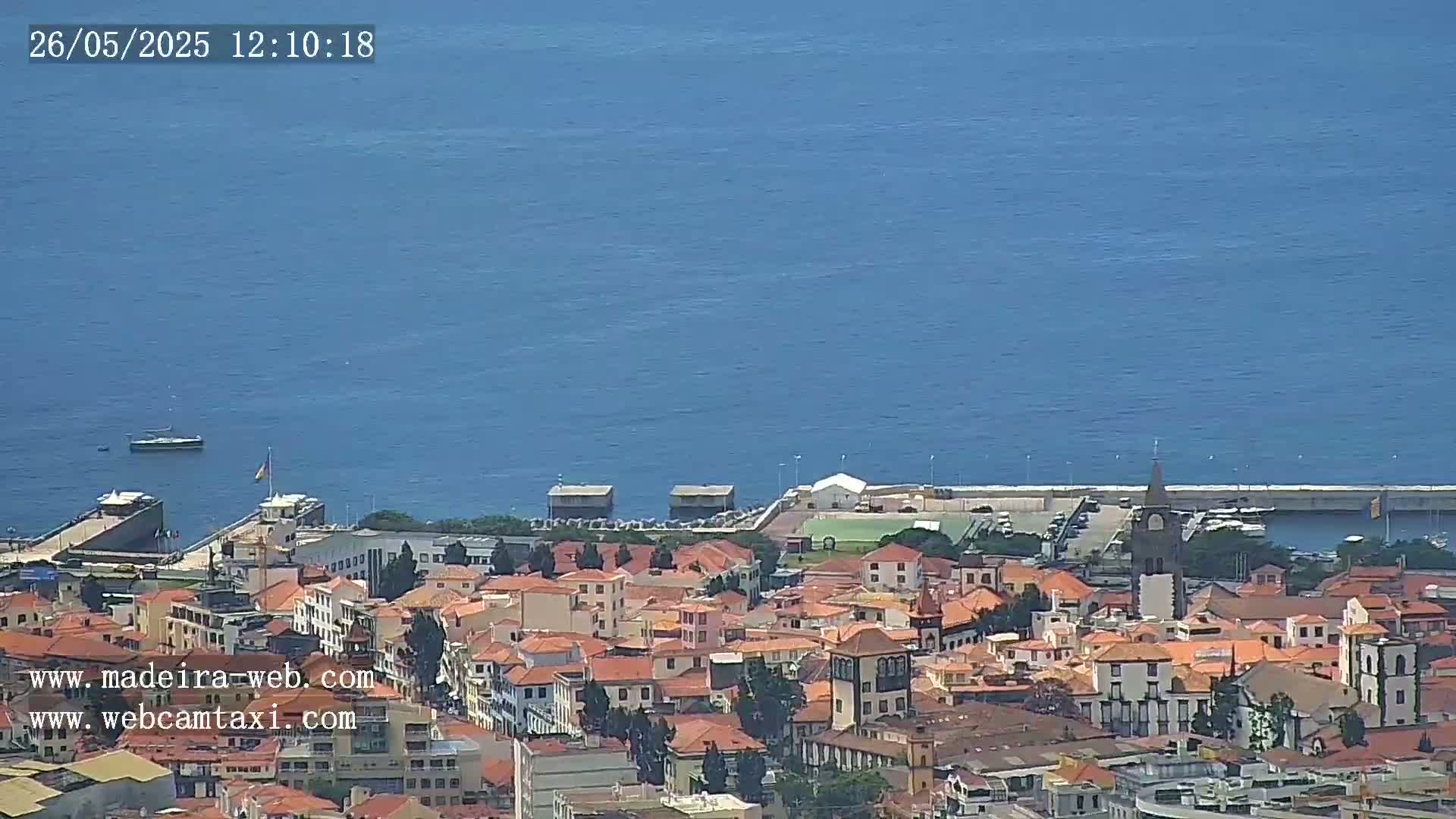 A coastal town with red-tiled roofs sits beside a calm blue ocean under a clear sky.