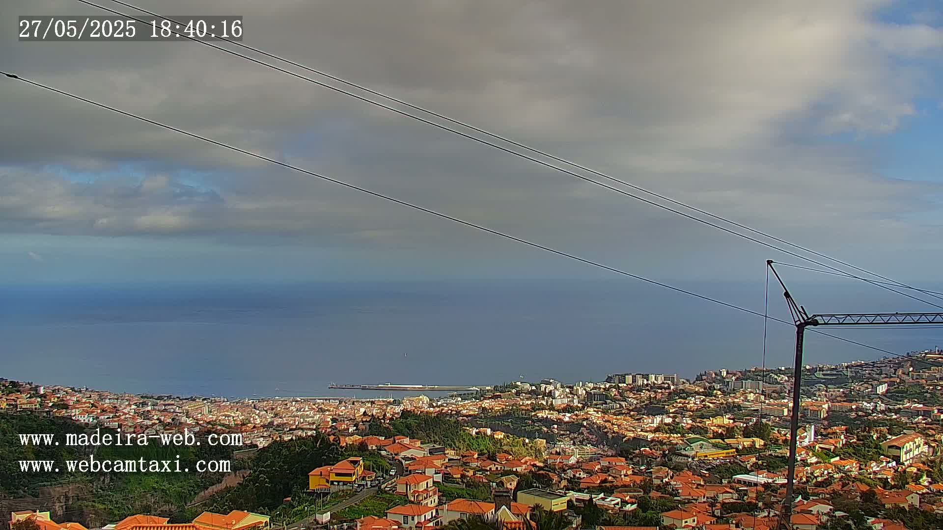 A coastal city with red-roofed buildings sprawls below a partly cloudy sky, overlooking a calm ocean.