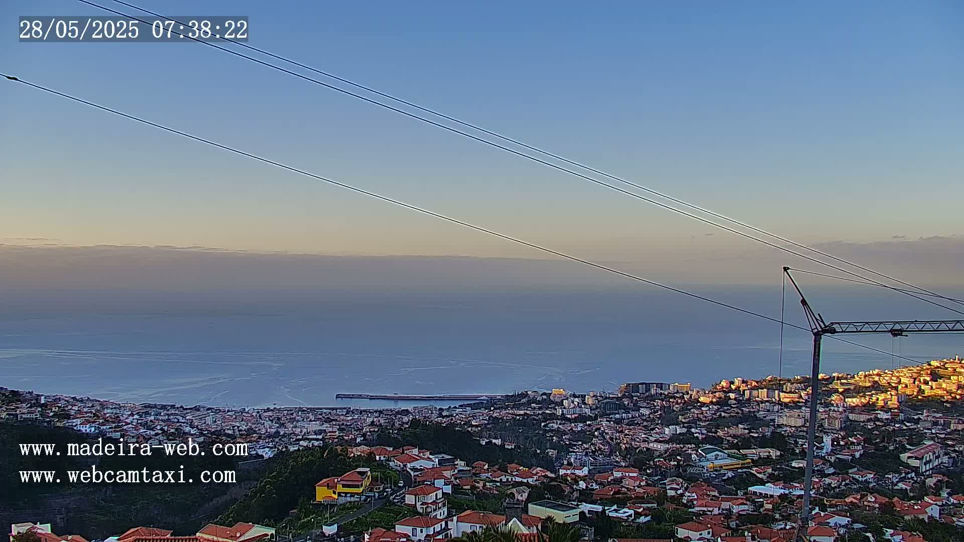 A coastal city at sunrise is seen from a hilltop, with calm ocean waters stretching to the horizon under a clear sky.