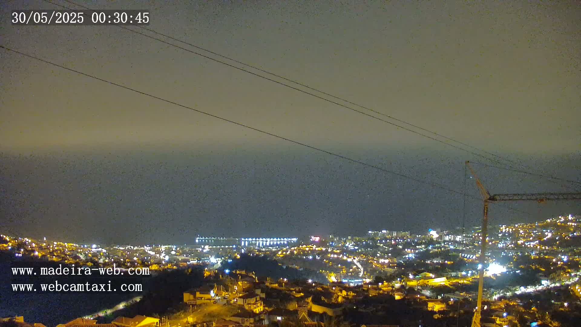 A nighttime, hazy view shows a coastal city with illuminated buildings, a crane, and power lines against a dark ocean under a muted sky.