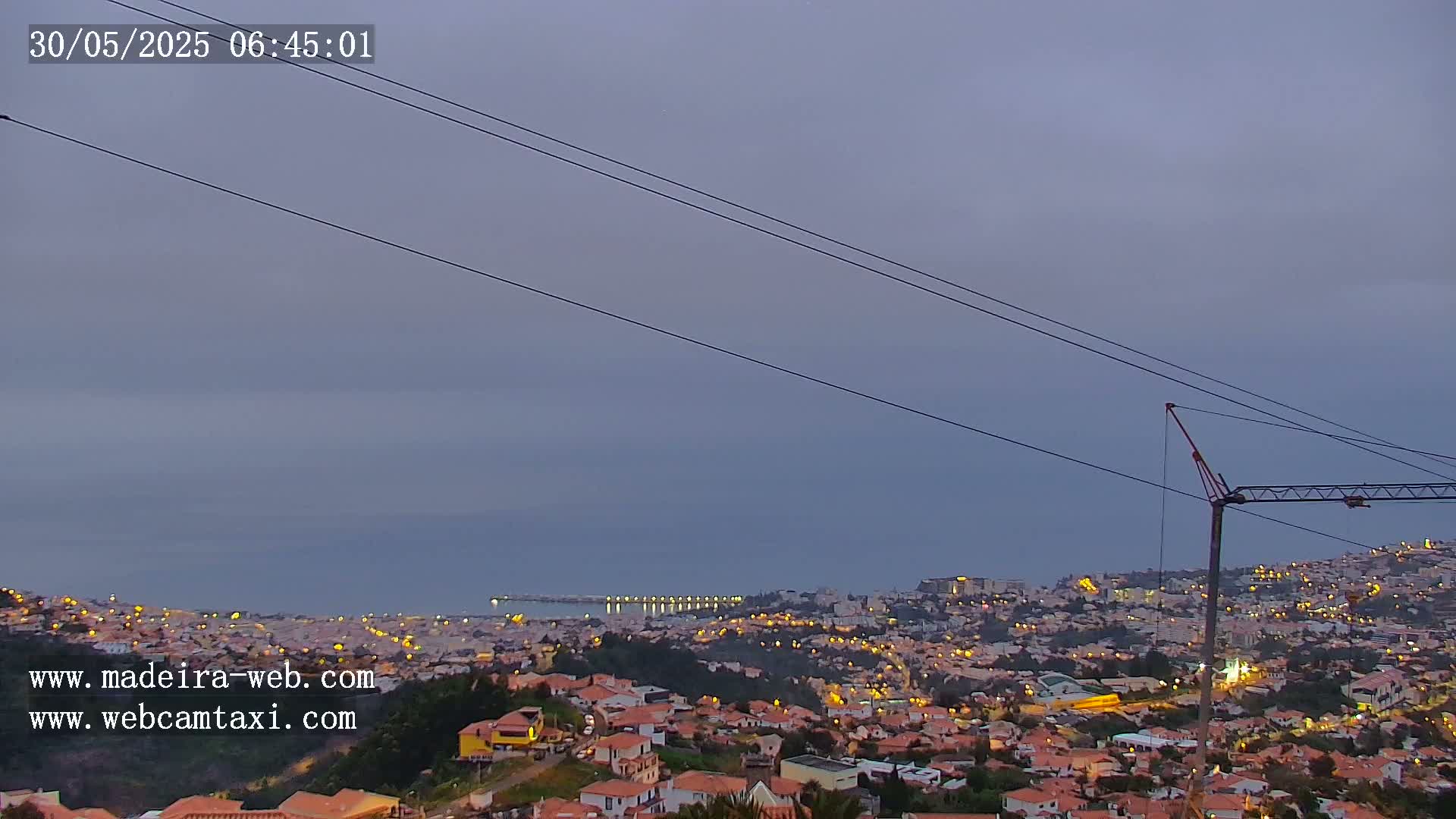 A twilight view overlooking a city illuminated with lights, a calm ocean, and a construction crane under a dusky sky.