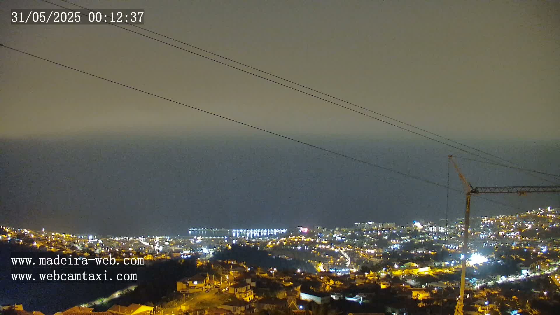 A nighttime view of a coastal city illuminated with lights, under a hazy sky, shows power lines in the foreground and a construction crane in the lower right.