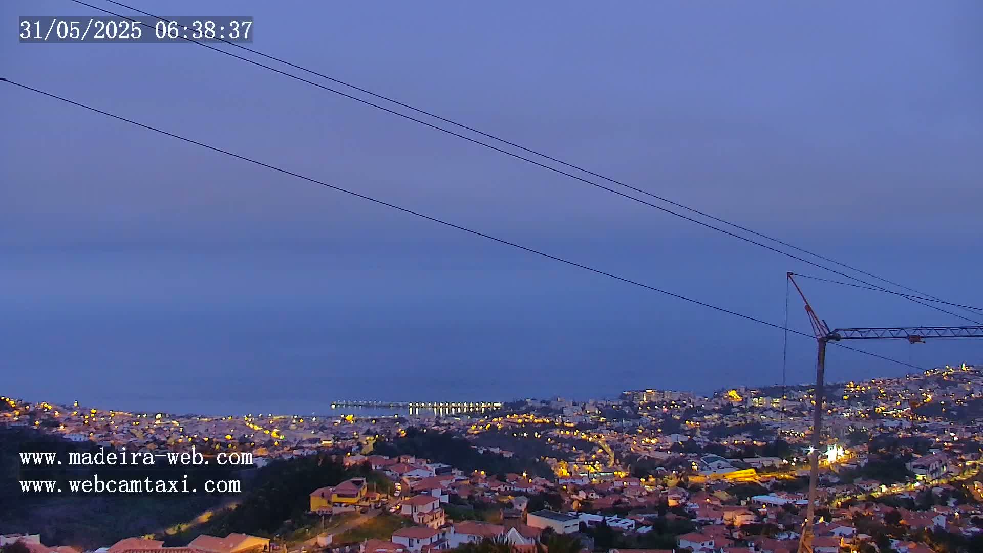 A twilight view shows a coastal town illuminated at night, with power lines and a crane in the foreground, overlooking a calm sea.