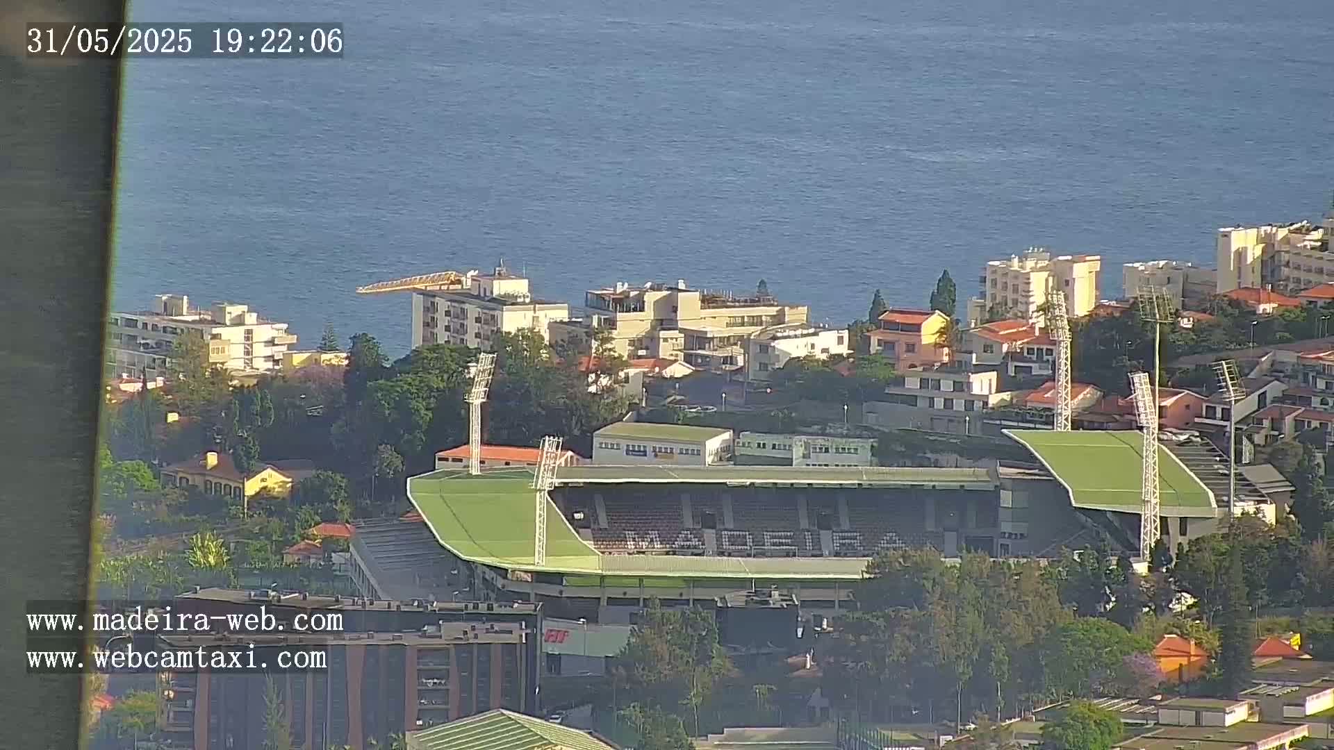A high-angle, long shot shows a stadium nestled among buildings overlooking a calm ocean on a sunny day.