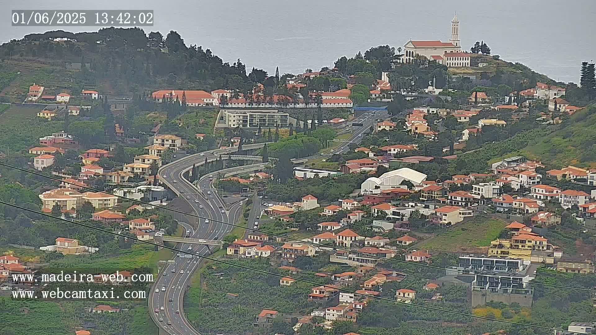 A hillside town with many low-rise buildings and a highway curving through it, overlooking a body of water under an overcast sky.