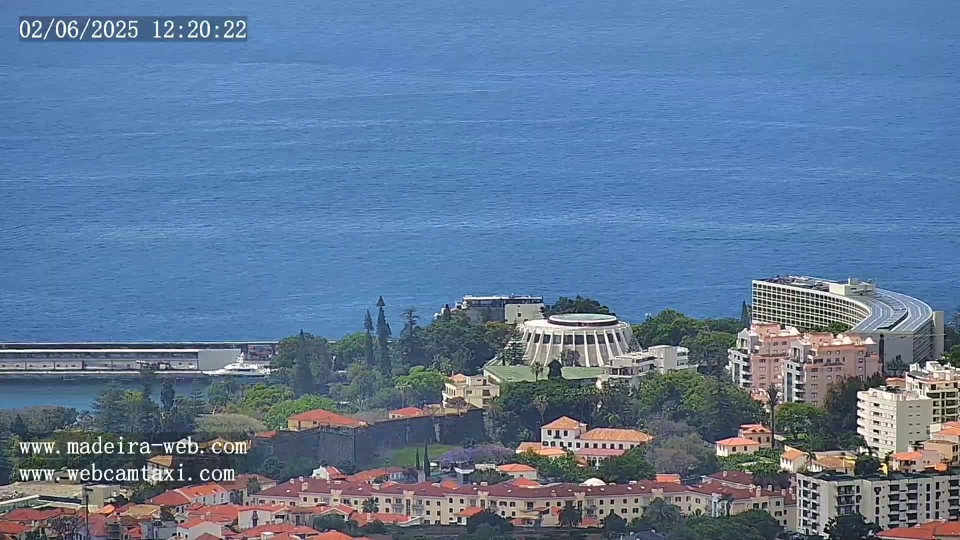 An aerial view shows a coastal town with buildings, green vegetation, and a large body of calm blue water under a clear sky.