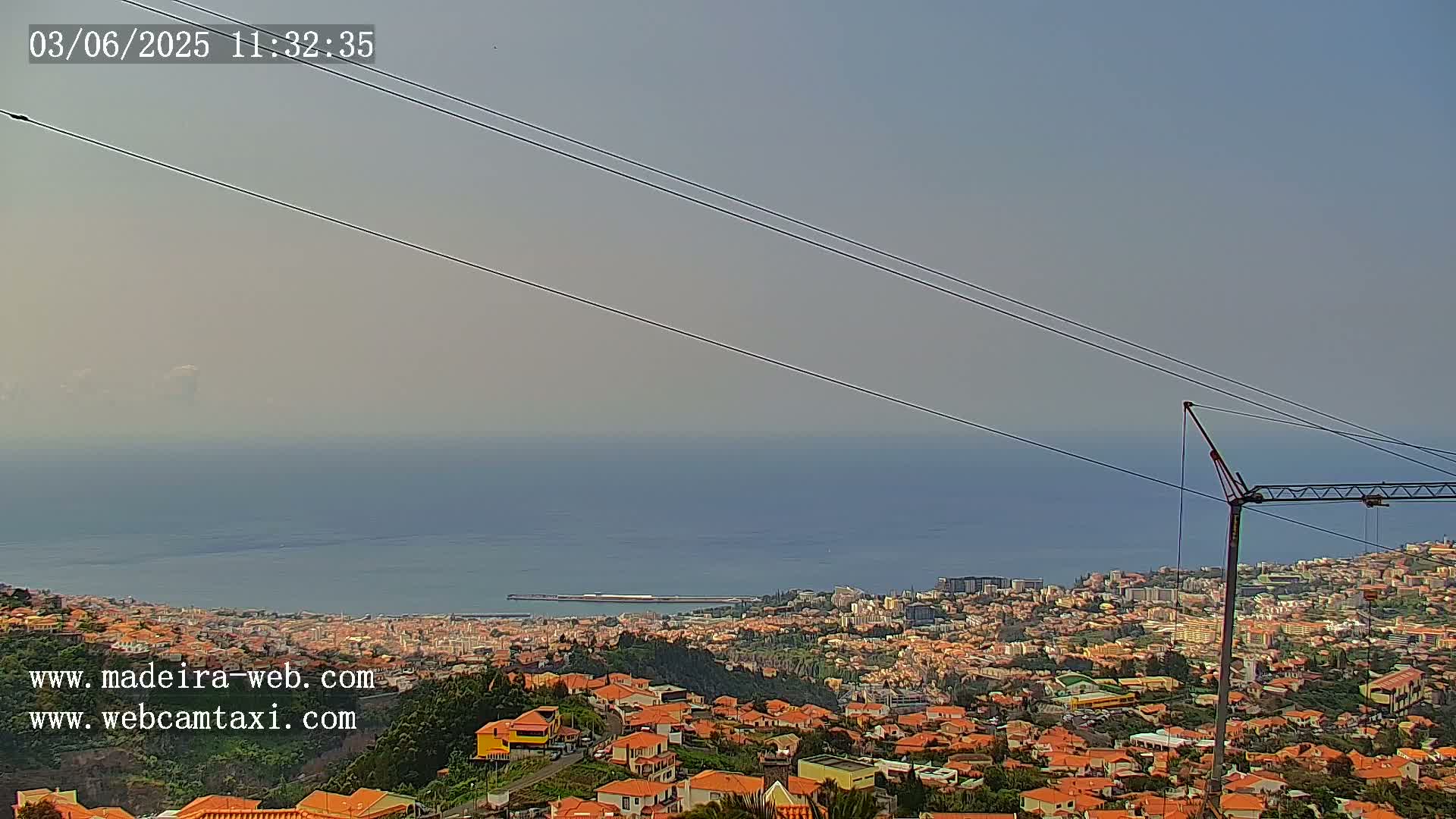A hazy, sunny day overlooks a coastal town with red-roofed houses, a body of water, and a construction crane in the foreground.