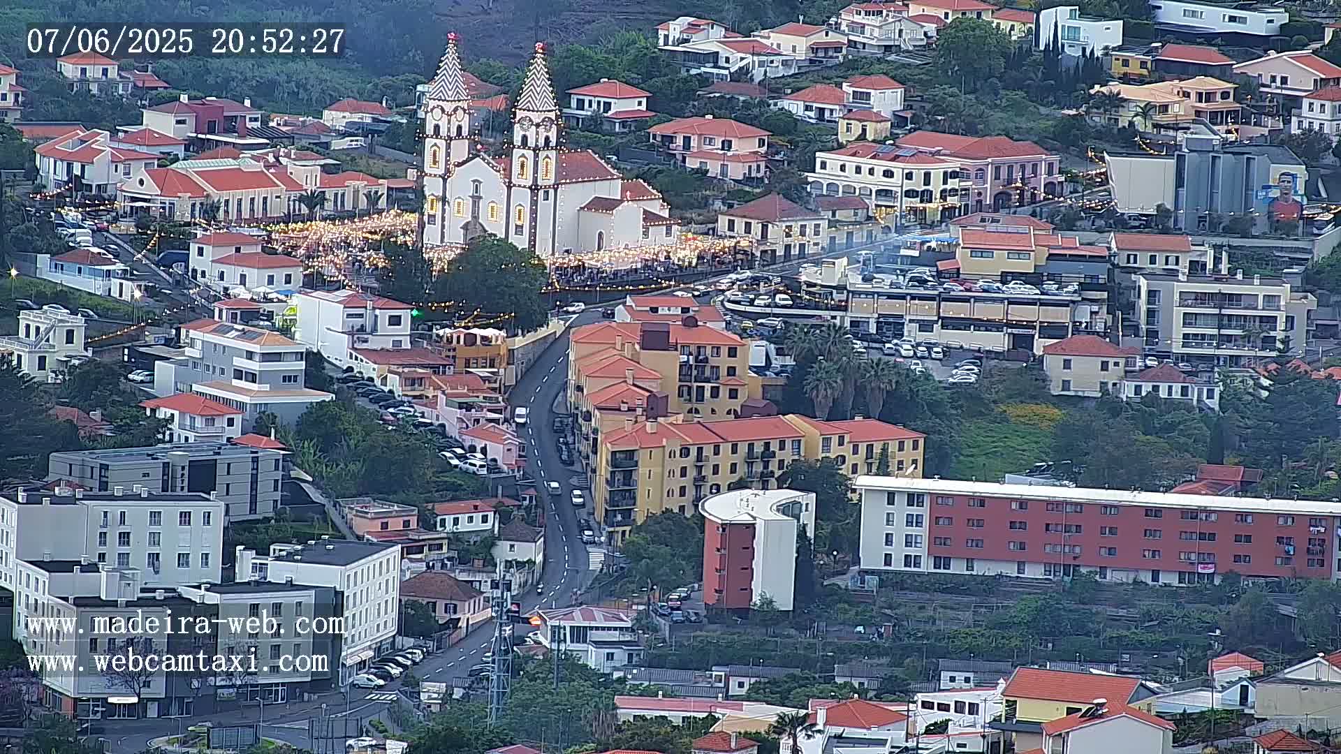 An aerial view shows a town at dusk, with a large white church illuminated, surrounded by buildings and streets filled with cars, under a mostly clear sky.