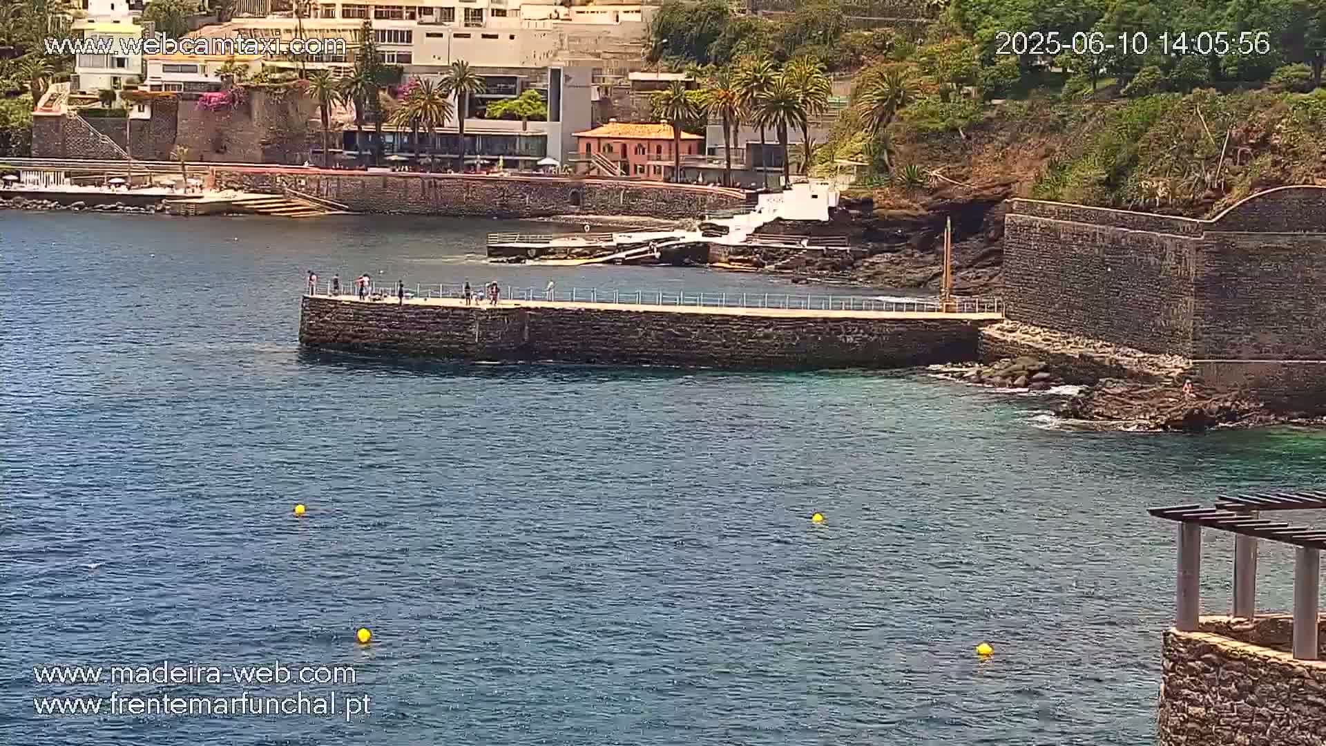 A calm, sunny day shows a stone pier extending into a dark blue body of water near a shoreline with buildings and palm trees.