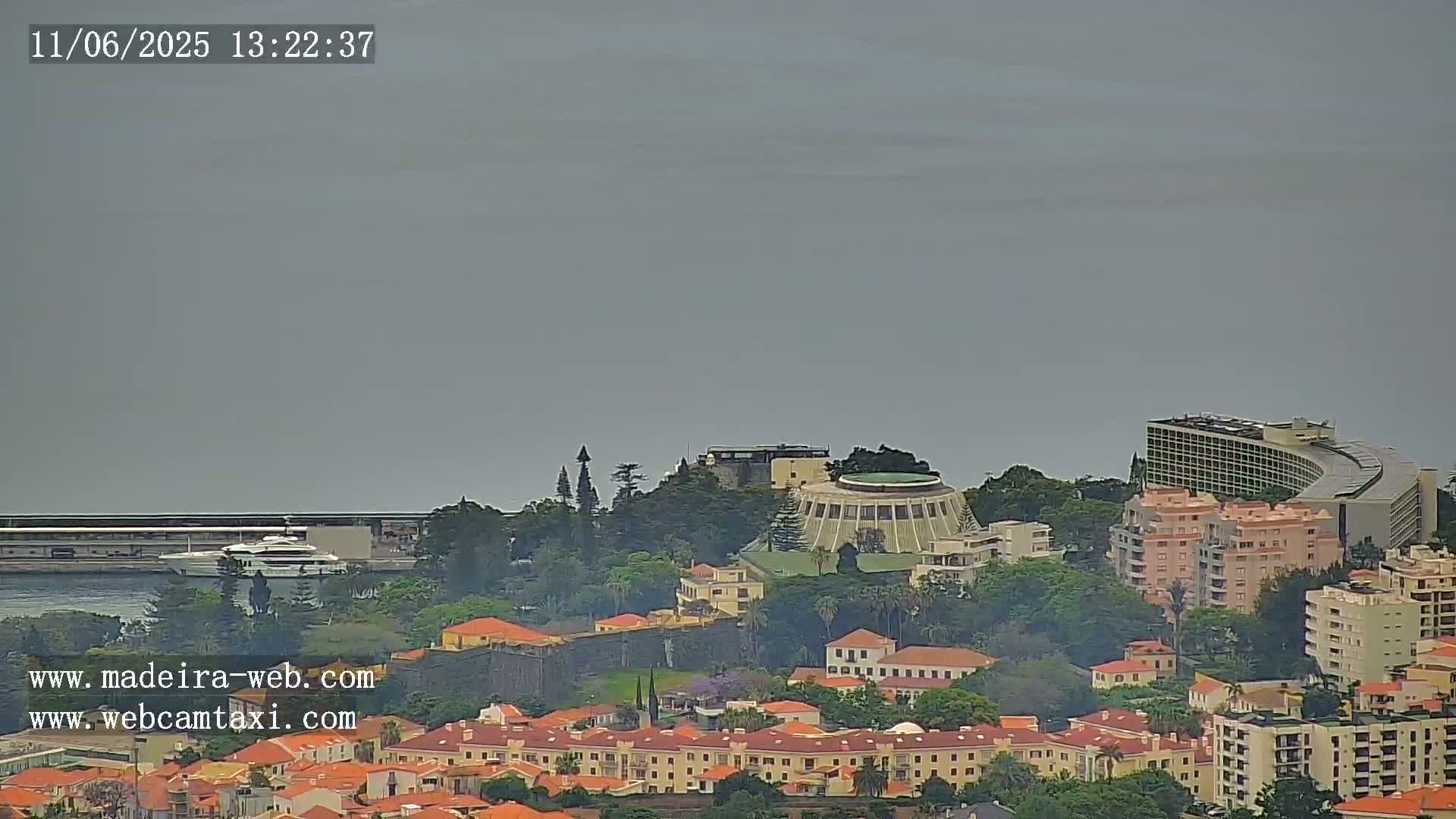 A hazy coastal city scene shows a harbor with a boat, a large building on a hilltop among other buildings, and many residential structures extending down to the water.