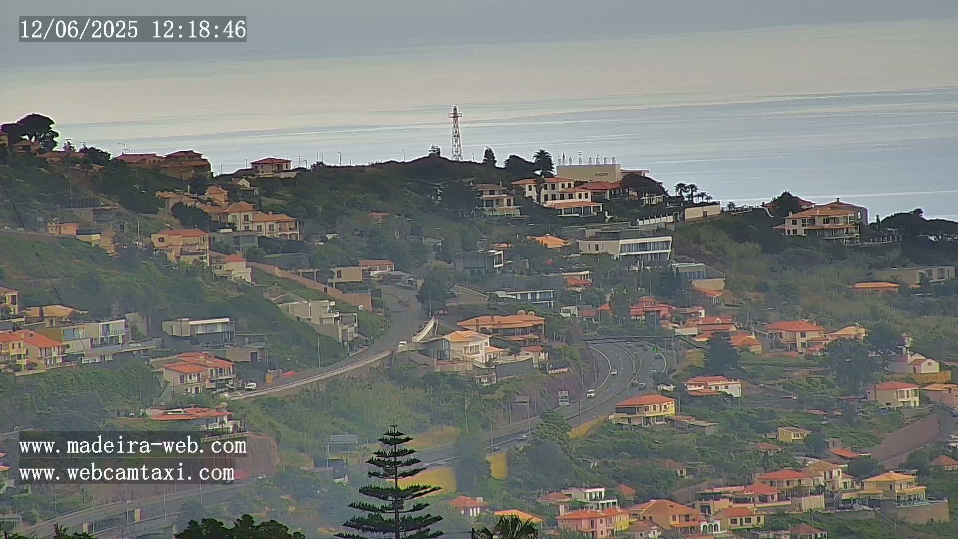 A hazy, overcast day reveals a coastal hillside town with numerous houses, a highway, and a calm ocean in the distance.