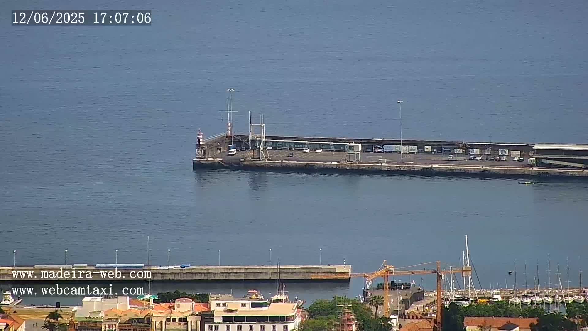 A long pier with several vehicles parked on it extends into a calm, blue body of water, with a smaller pier and a marina visible in the foreground.