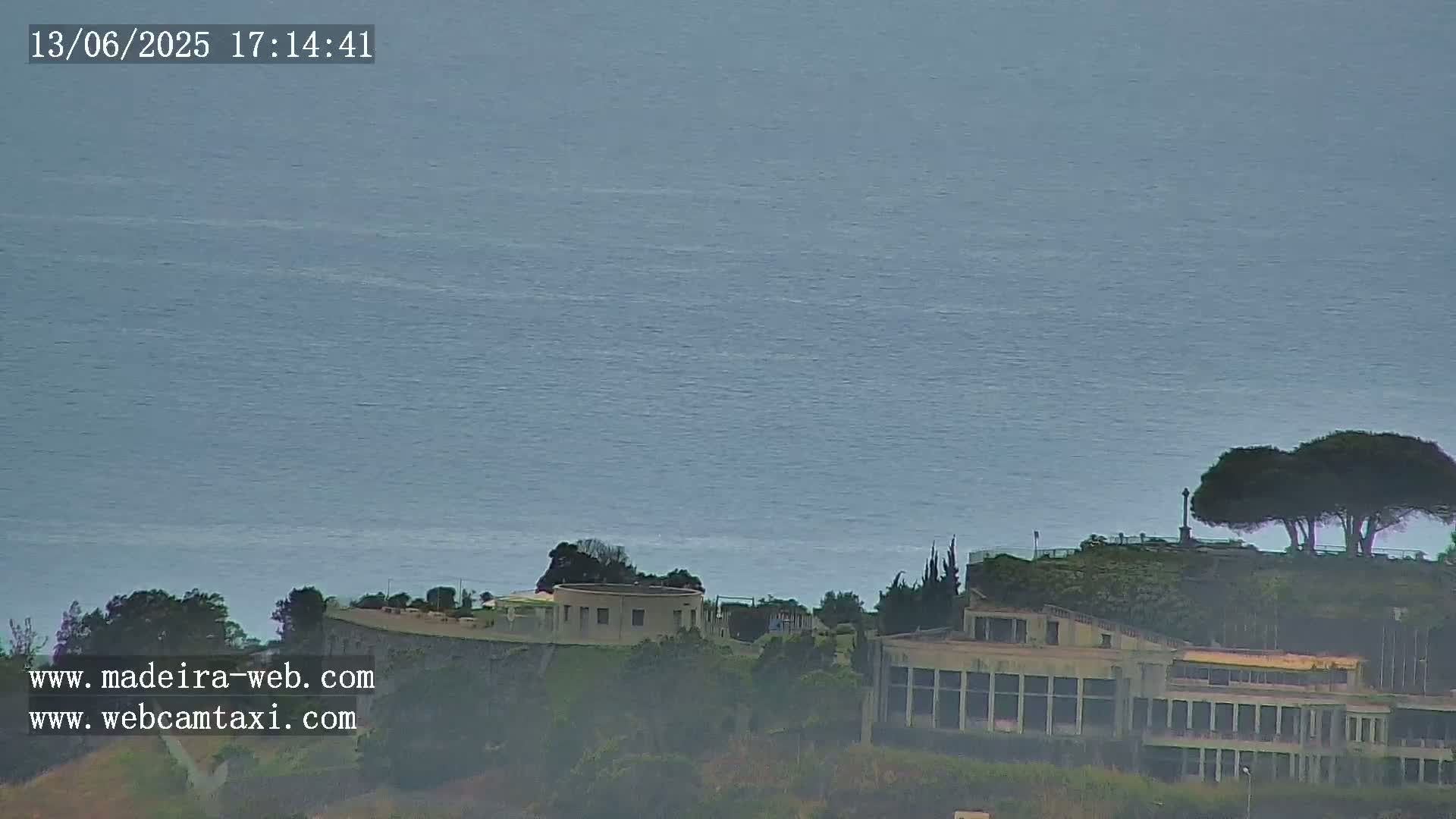A calm, gray ocean stretches to the horizon, overlooking a hillside with buildings and several dark green trees under a hazy sky.