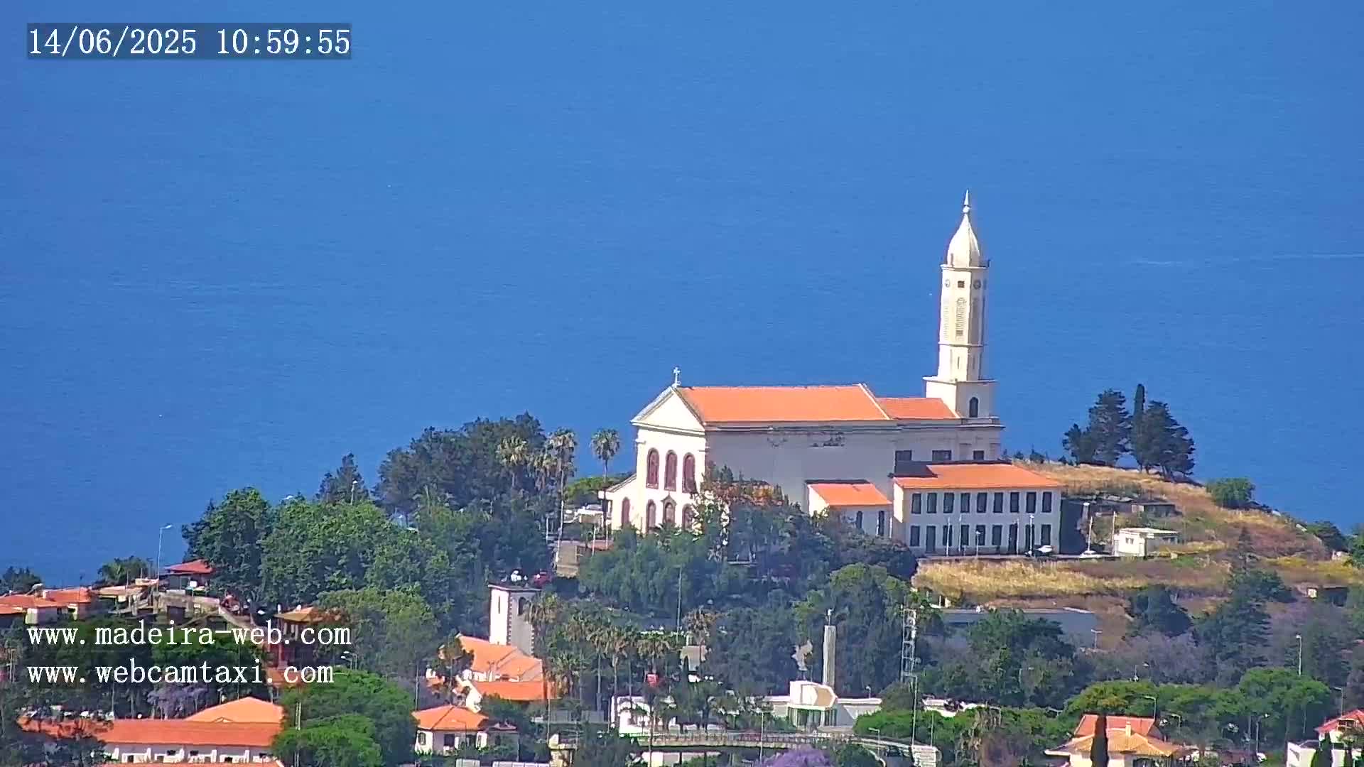 A white church with an orange roof sits atop a hill overlooking a calm blue ocean under a clear blue sky.