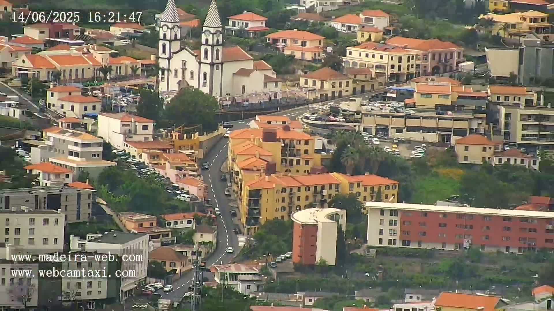 An aerial view shows a town with many orange-roofed buildings, a white church with two towers, and a winding road, under somewhat hazy conditions.