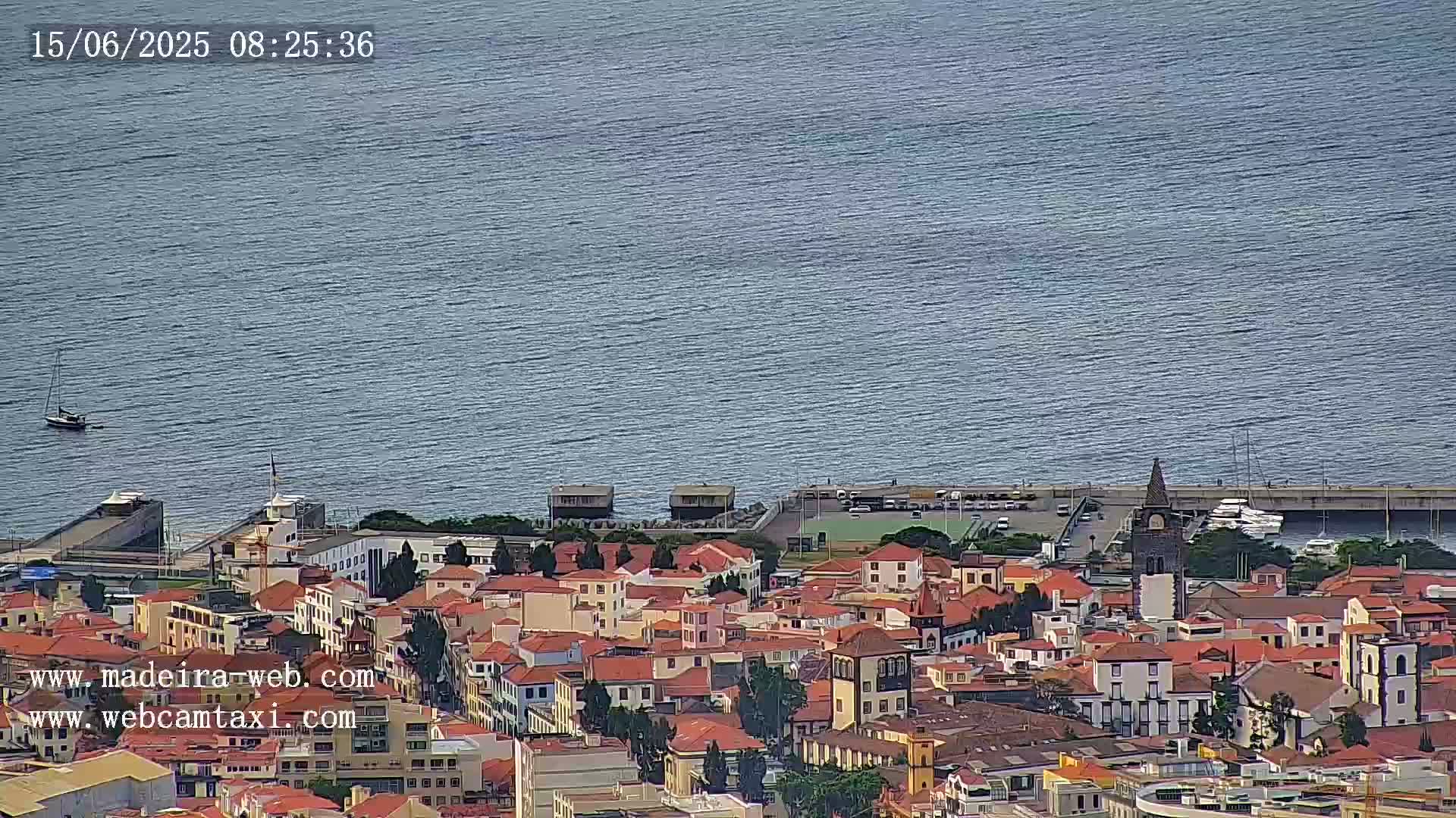 A coastal town with red-roofed buildings is situated beside a calm body of water with a few boats visible.