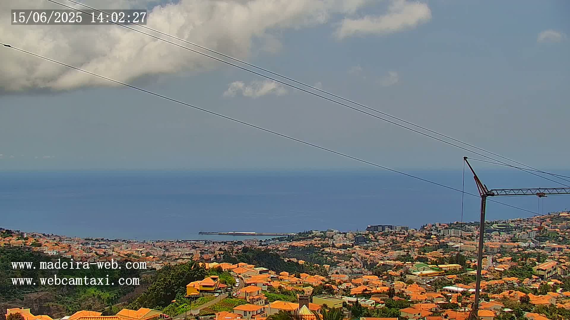 A partly cloudy day overlooks a coastal town with orange-roofed houses, a calm ocean, and a construction crane.