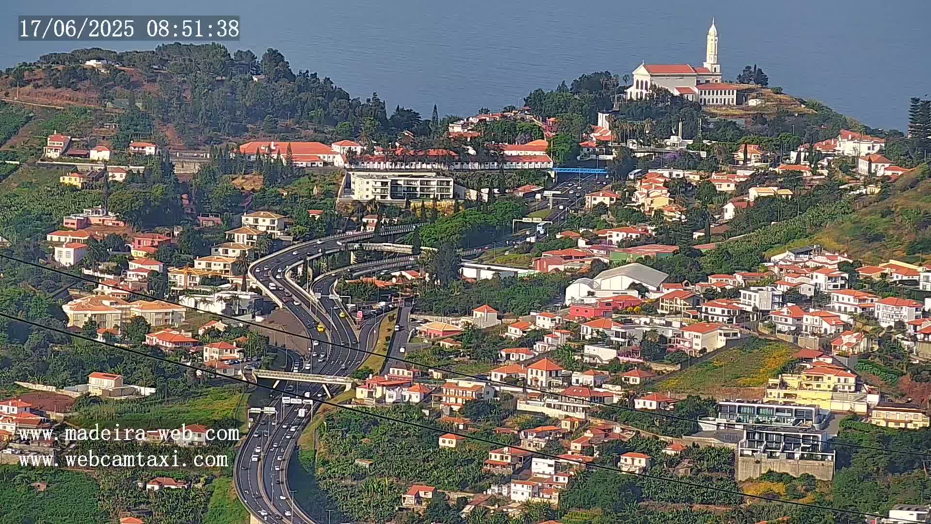 A hillside town with many low-rise buildings, a highway curving through it, and a church on a hilltop overlooking a calm ocean under a clear sky.